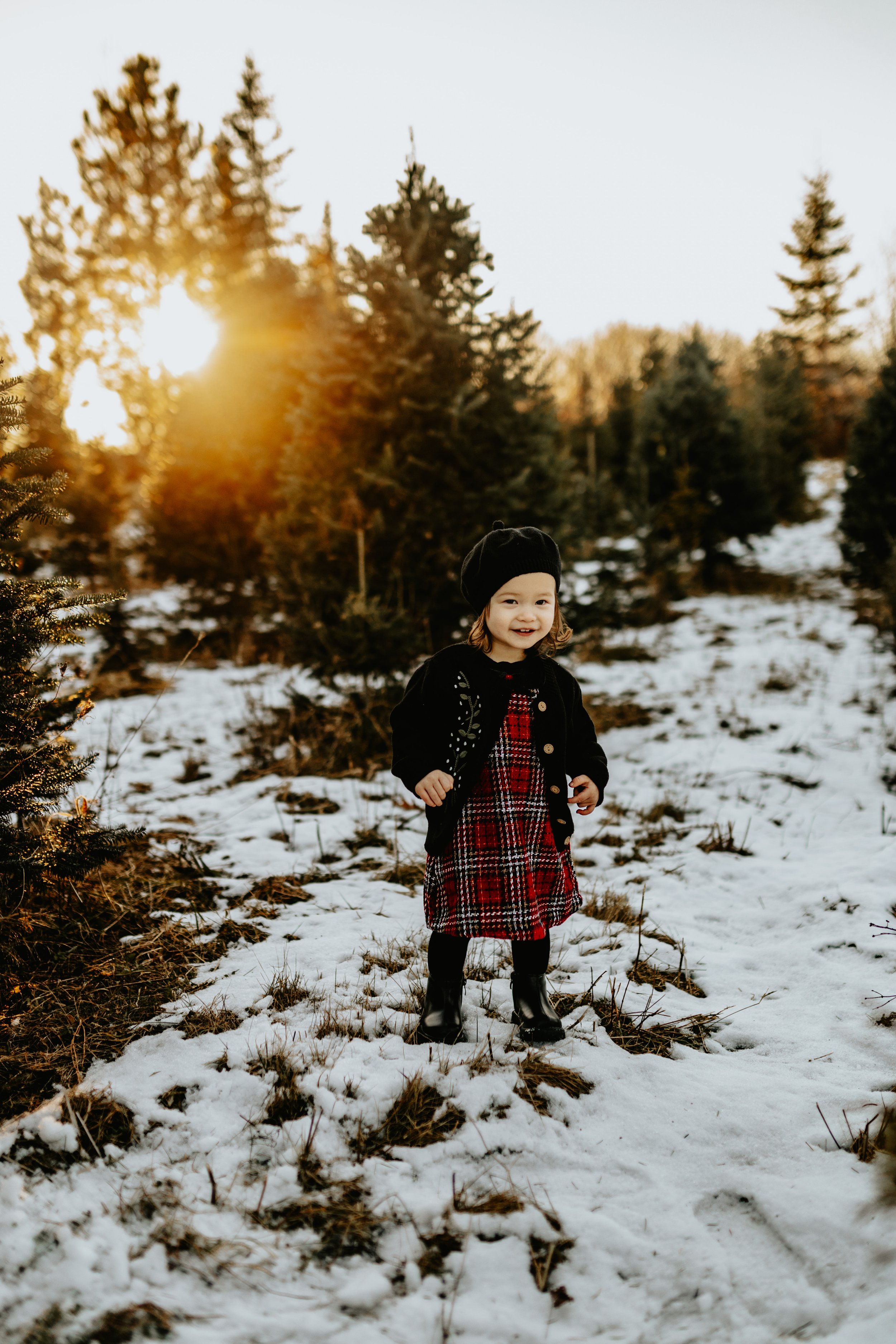Young child in winter clothing standing on snow-covered ground with pine trees and sunset in the background.