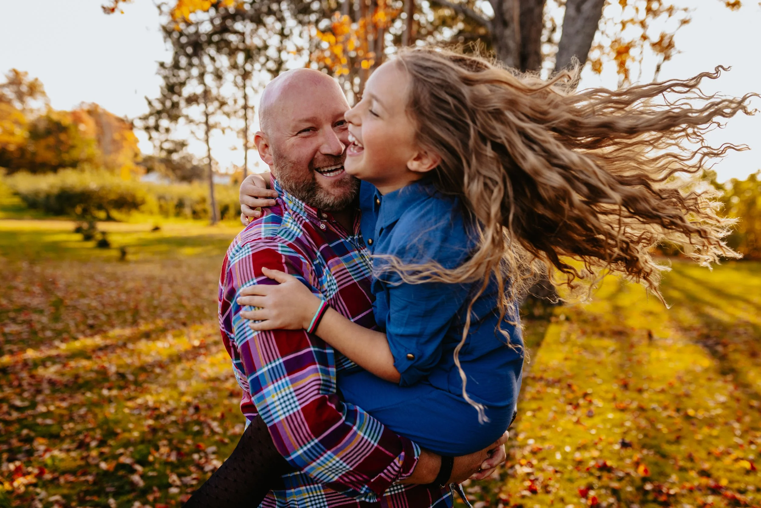 A man holding a young girl in a park during autumn, they are smiling and laughing with trees and fallen leaves in the background.