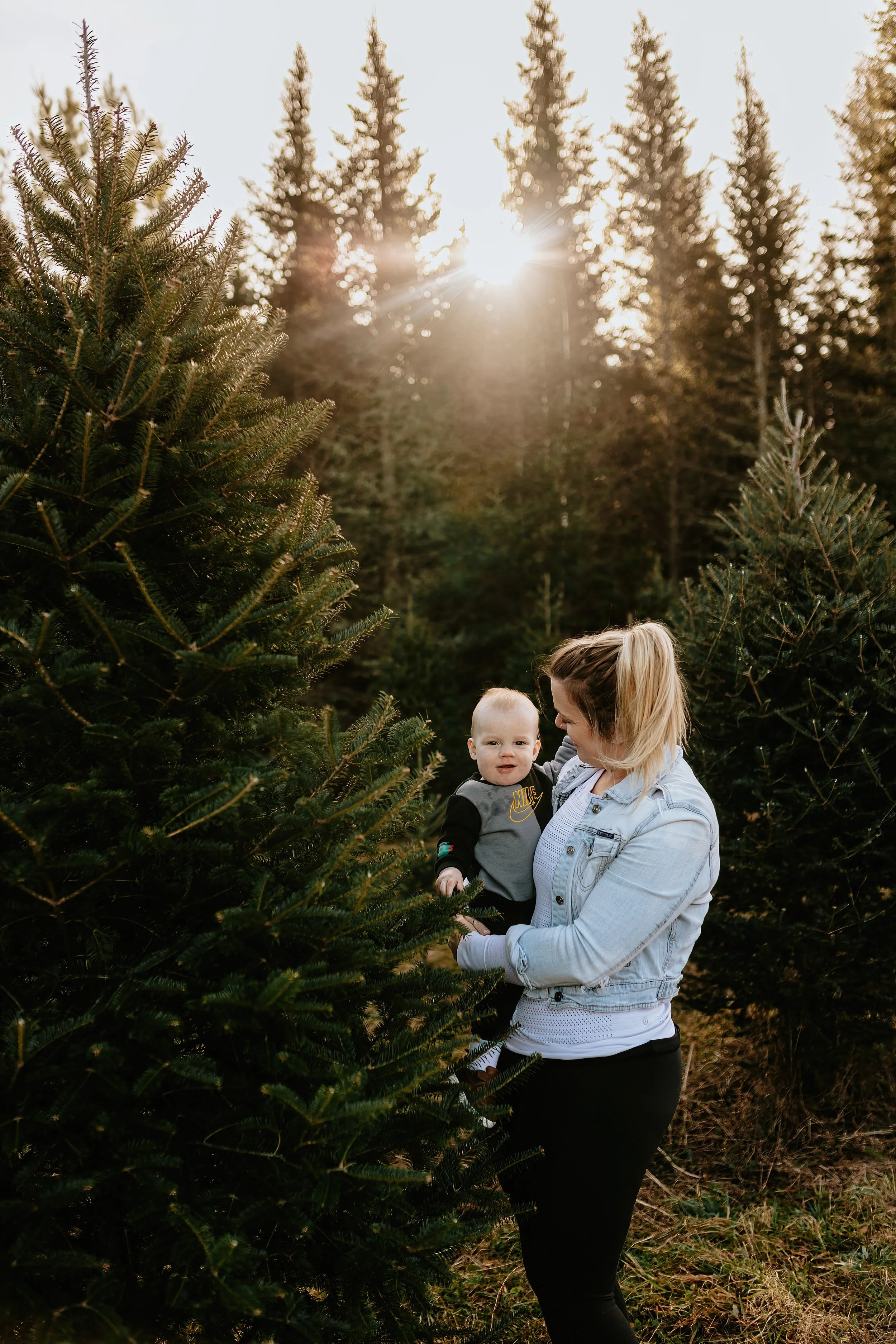 Woman holding a baby standing near Christmas trees, with sunlight filtering through the trees.