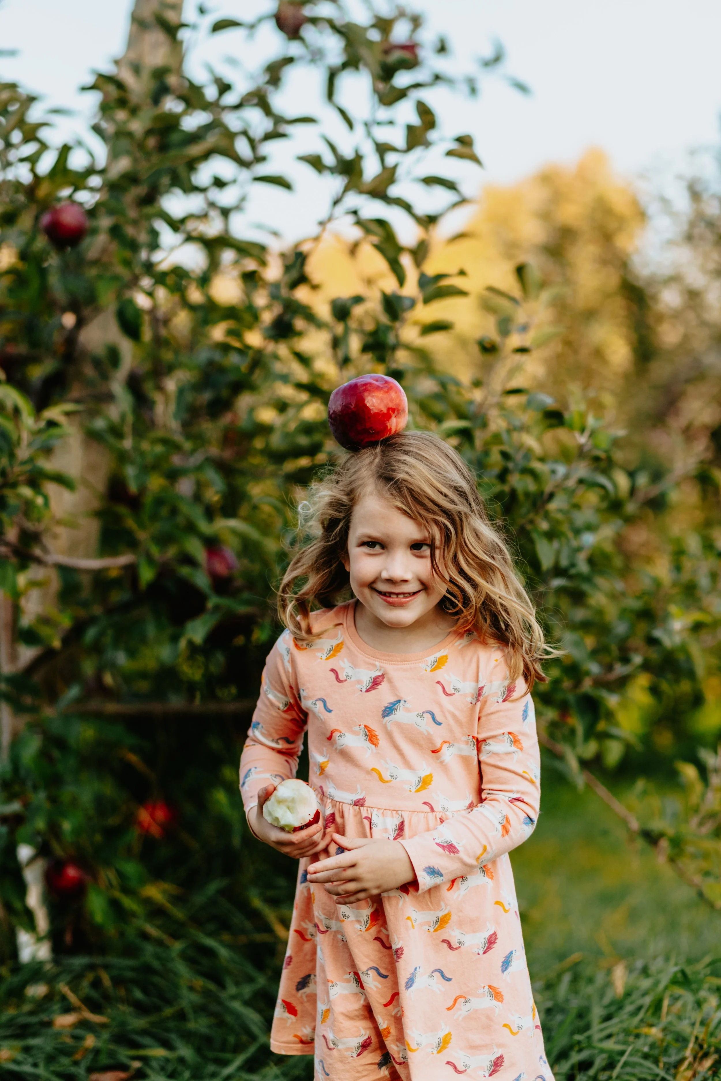 A young girl with wavy blonde hair standing outdoors in an apple orchard, balancing an apple on her head while holding another apple in her hand, smiling.