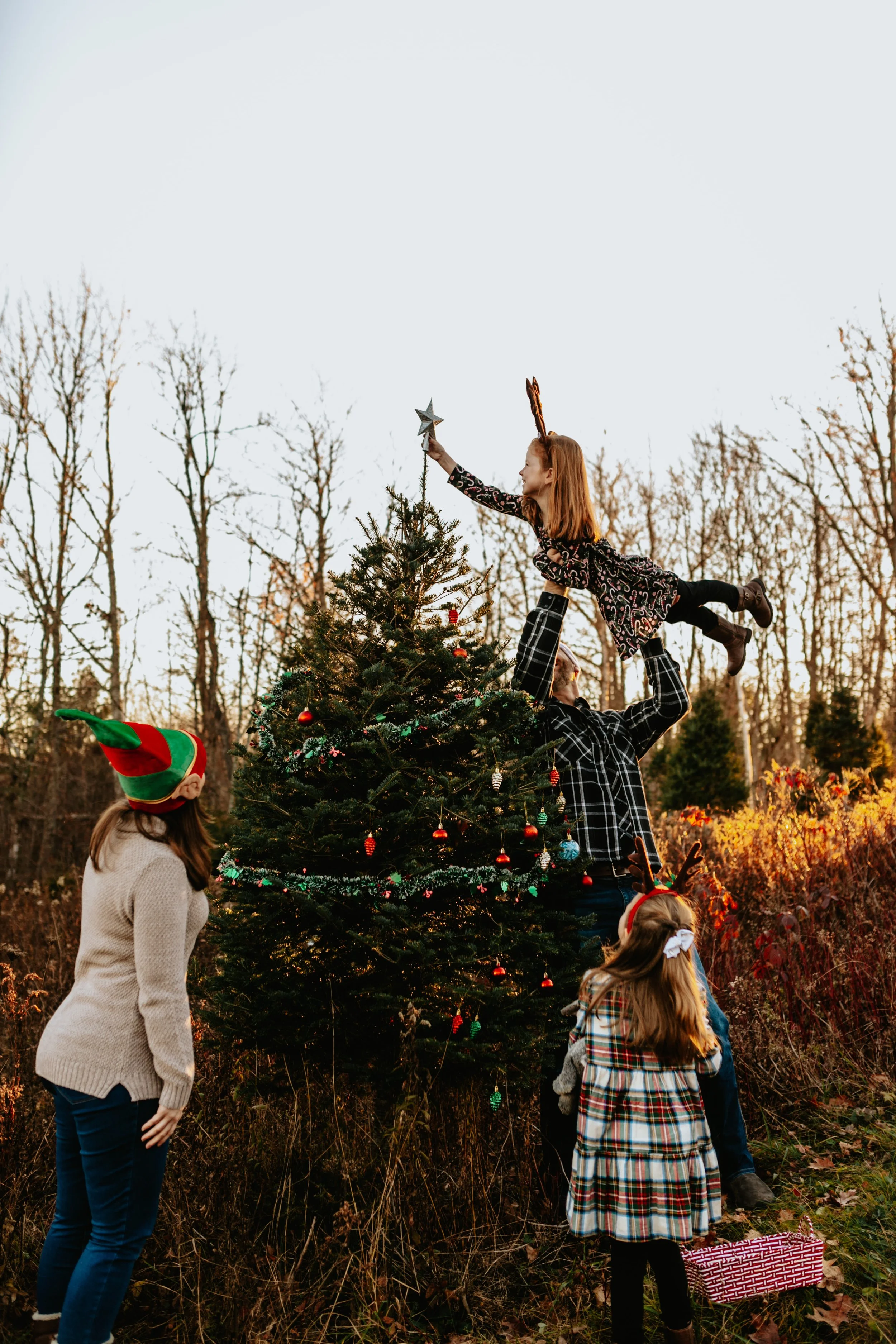 A family stands around a decorated evergreen tree on a tree farm. The dad lifts his daughter up to place the star on top.