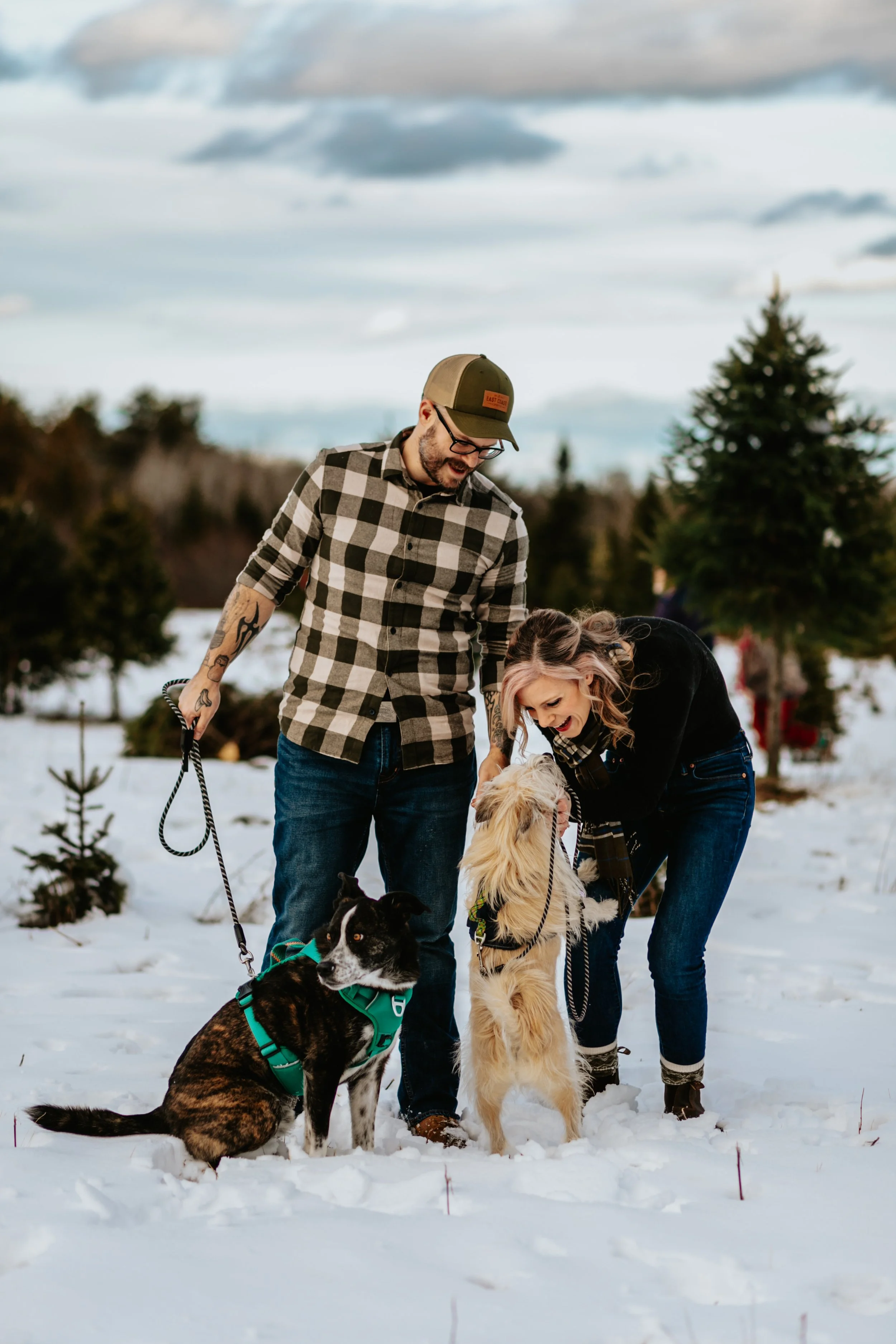 A man and woman are standing outdoors in snowy terrain with two dogs, smiling and interacting with the dogs.