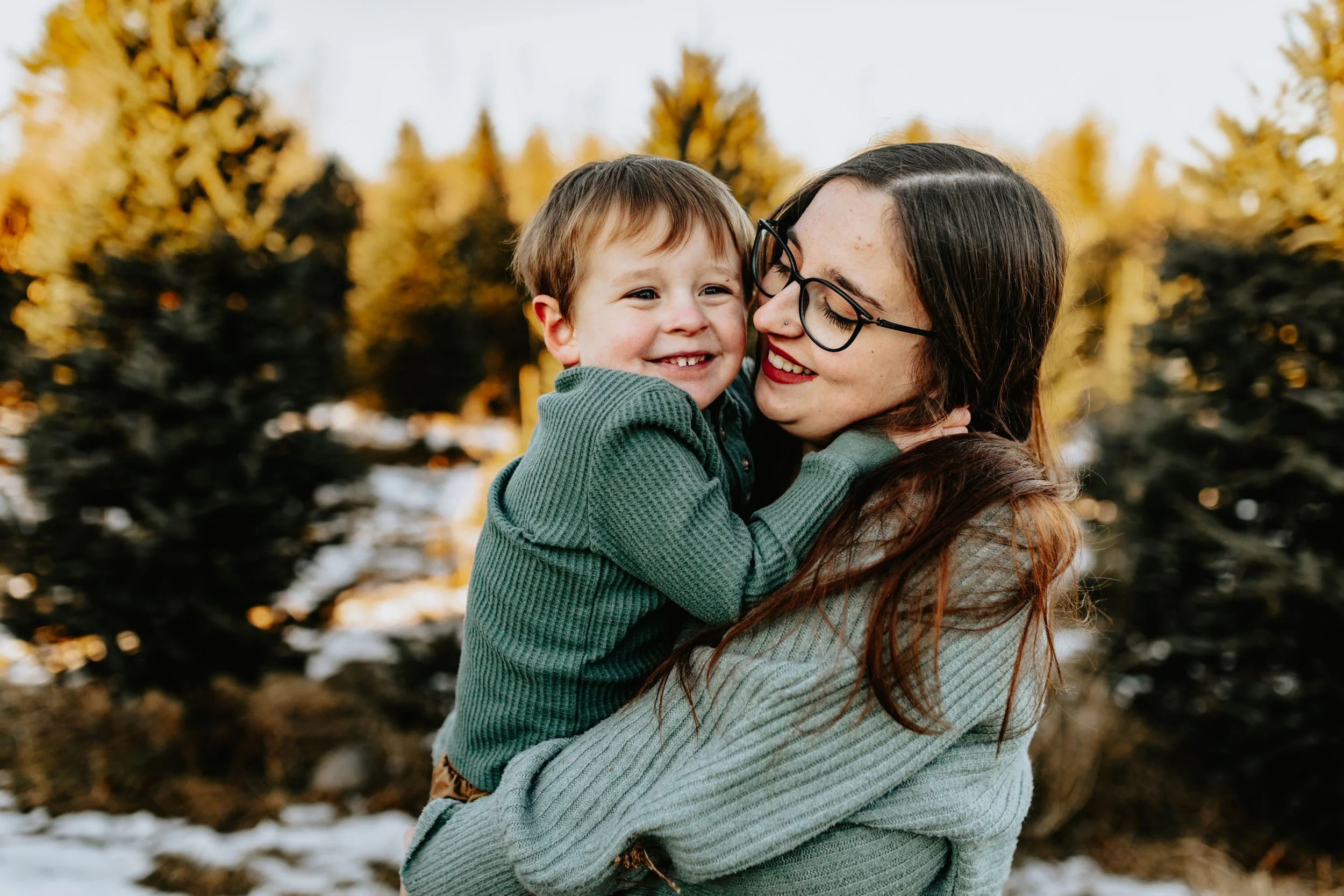 A woman holding a smiling child in a winter forest setting.