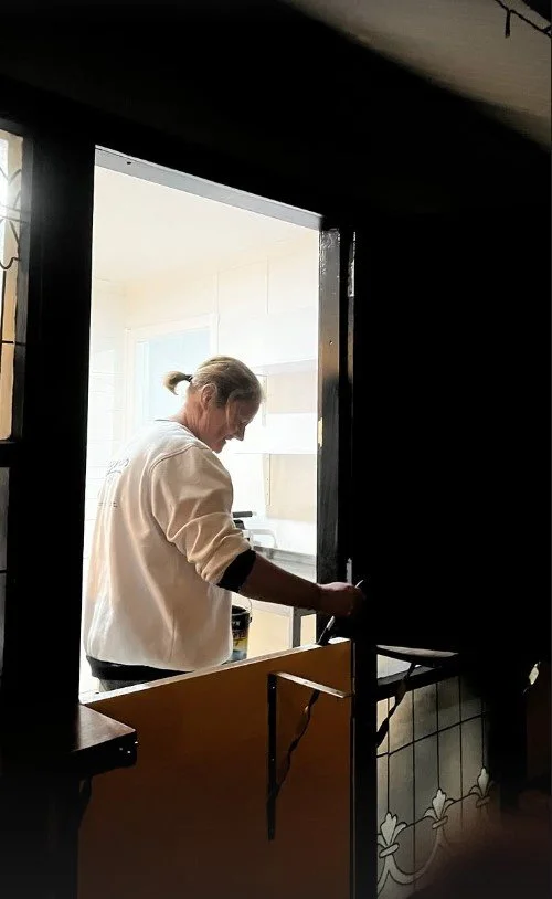Woman with a ponytail wearing a light-colored sweatshirt, standing in a bright kitchen, seen through a window or doorway from a darker room.