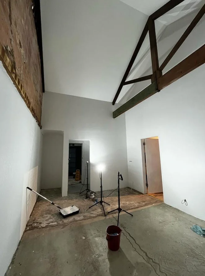 An unfinished room undergoing renovation or filming, with a floor cleaning machine, a mop, two lights on tripods, a red bucket, and visible exposed brick and beams.