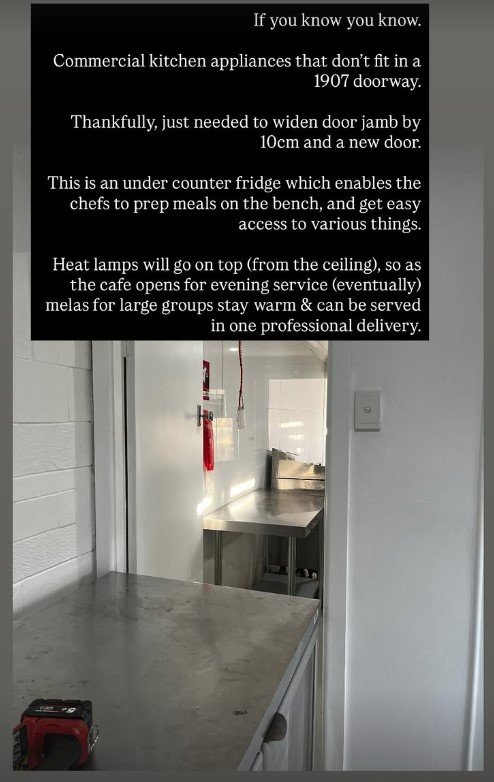 Interior view of a commercial kitchen with a counter, a small fridge underneath, and a door in the background. An informational sign explains upcoming modifications to the kitchen, including widening the doorjamb and installing heat lamps on the ceiling for evening service.