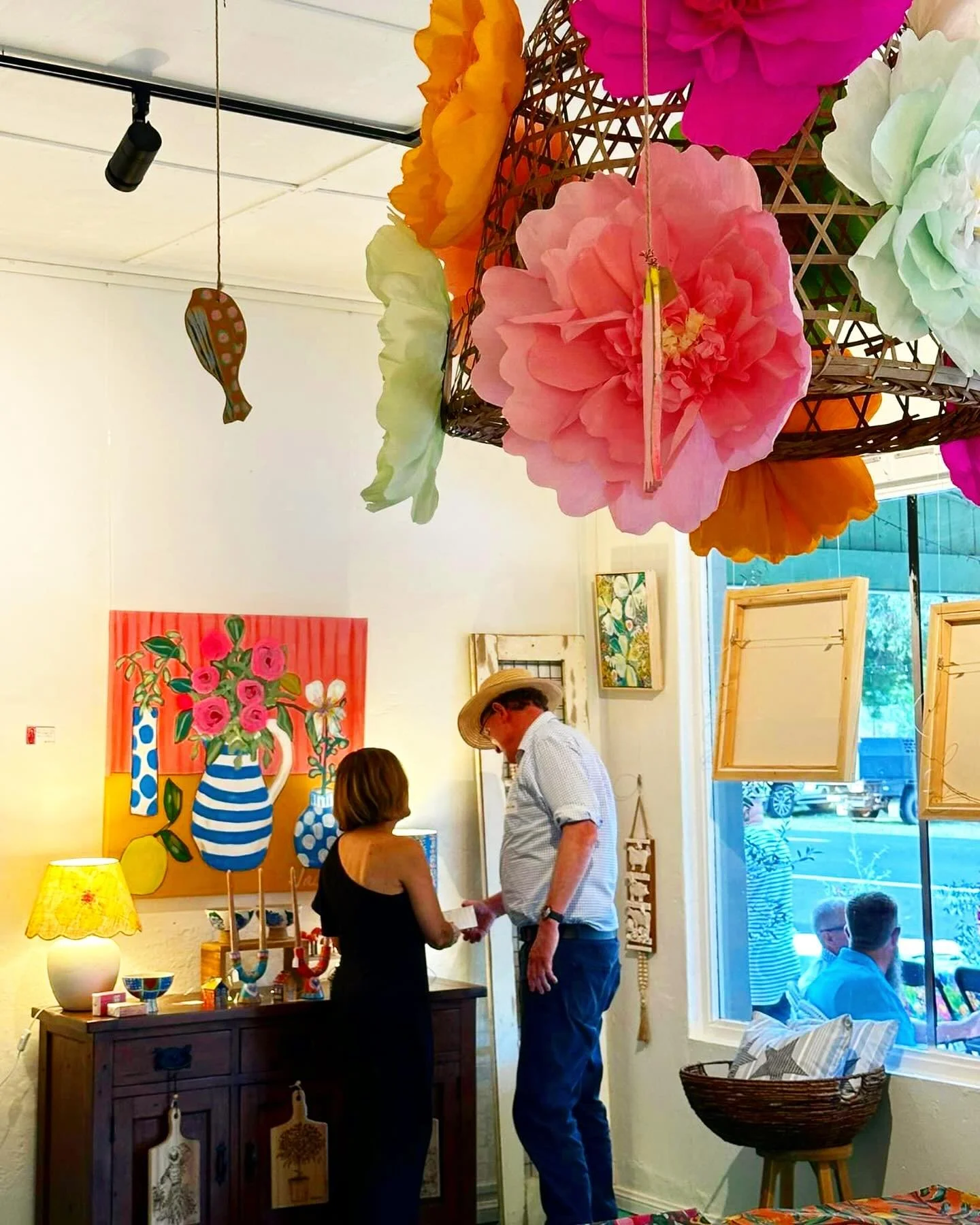 Two people are standing and talking inside an art gallery decorated with colorful paper flowers hanging from the ceiling, art paintings on the walls, and various decorative items on a wooden dresser near a large window with a view outside.
