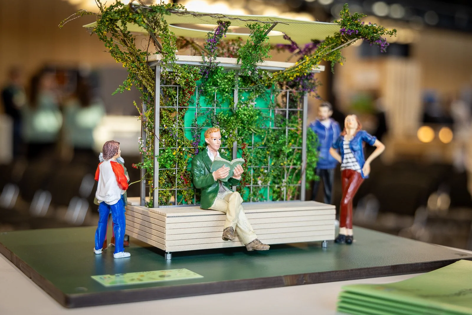 Modell eines mobilen Gartens mit Figuren: eine sitzende Figur liest, drei weitere Figuren stehen daneben, im Hintergrund sind Lichter und unscharfe Objekte.