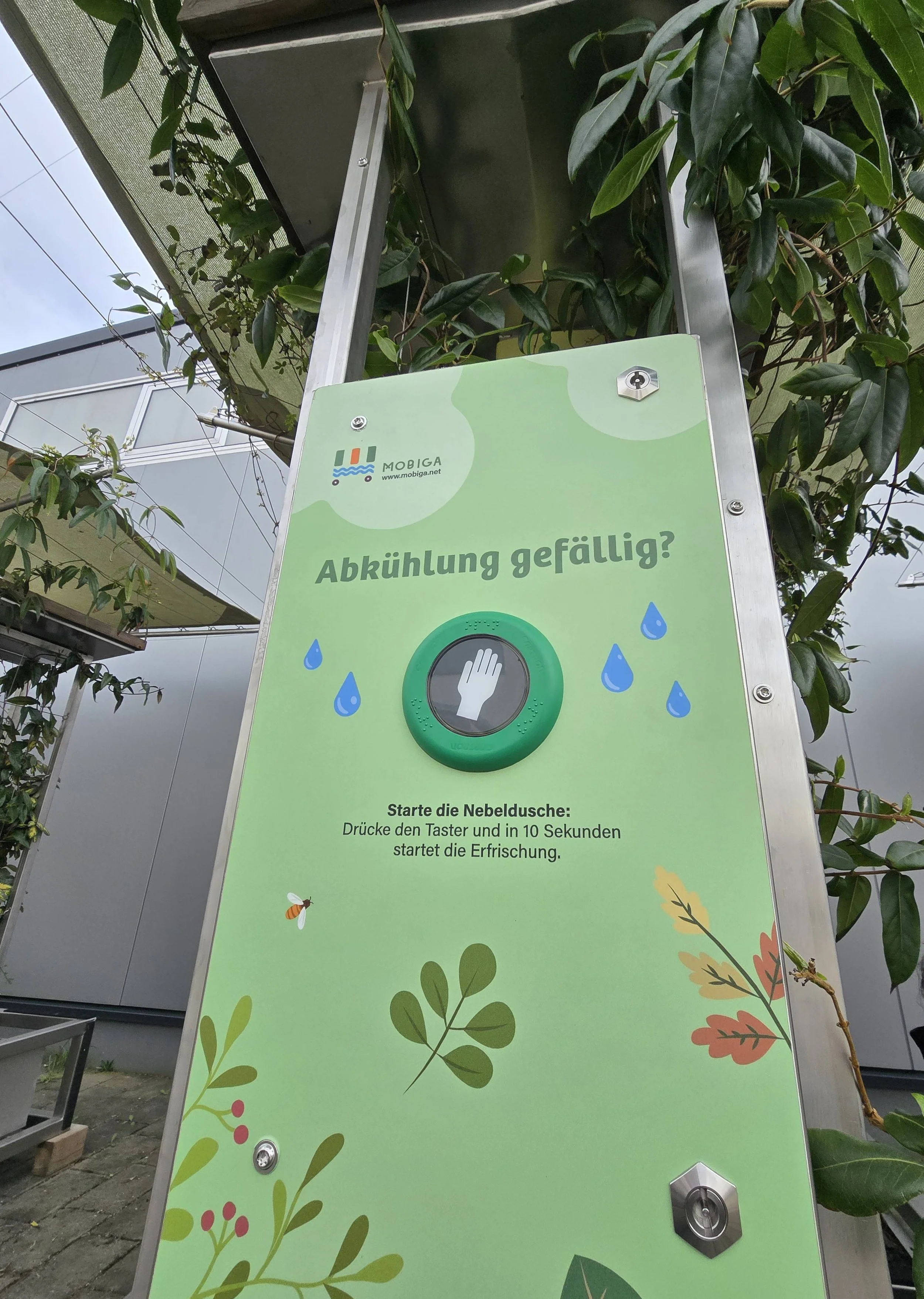 Hinweistafel mit der Frage 'Abkühlung gefällig?' und einem grünen Knopf, der die Nebeldusche startet, mit Anweisungen zur Bedienung in Deutsch, um die Erfrischung zu aktivieren.