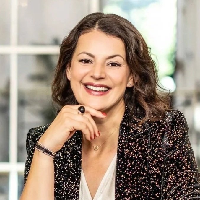 A woman with brown hair smiling, wearing a red blazer, standing indoors in front of a blurred background with shelves and greenery.