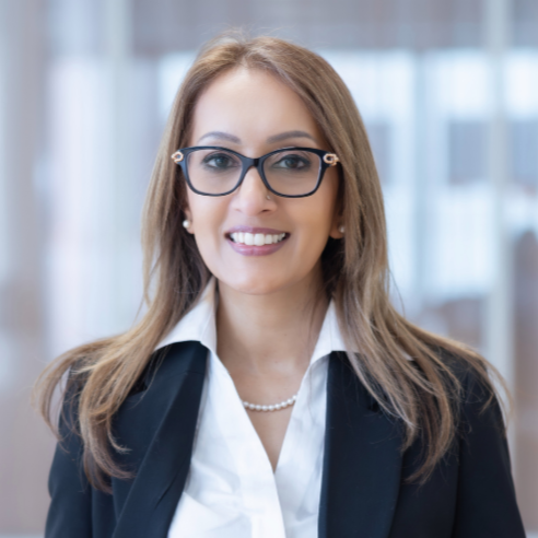 A smiling woman with long, wavy, reddish-brown hair, wearing a dark blazer and a small necklace, standing against a plain white background.