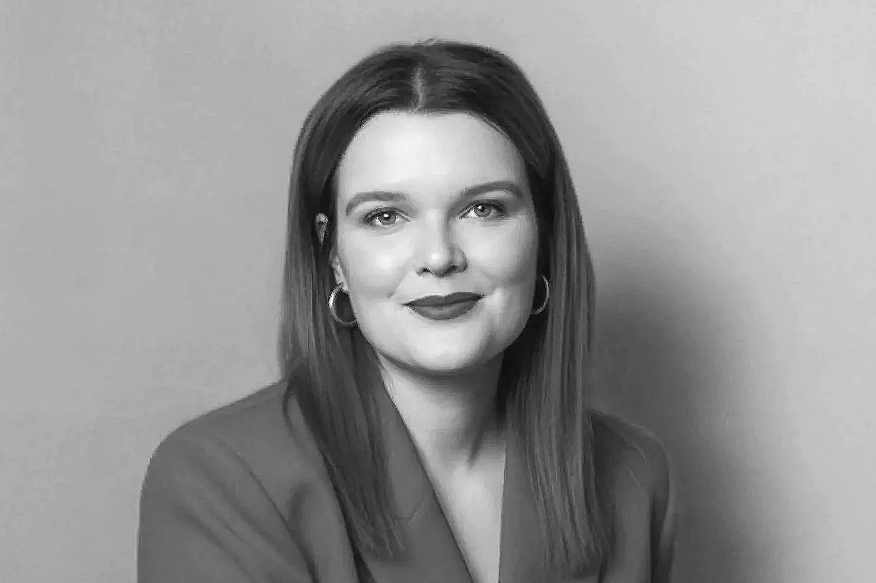 Schwarz-Weiß-Fotografie einer Frau mit dunklen, geraden Haaren, lächelnd, in dunkler Kleidung und Ohrringen vor einer neutralen Wand.