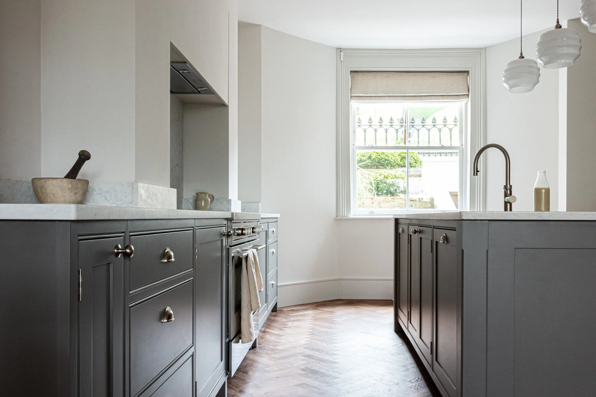 Looking between a row of grey painted shaker style bespoke kitchen cabinets towards a window