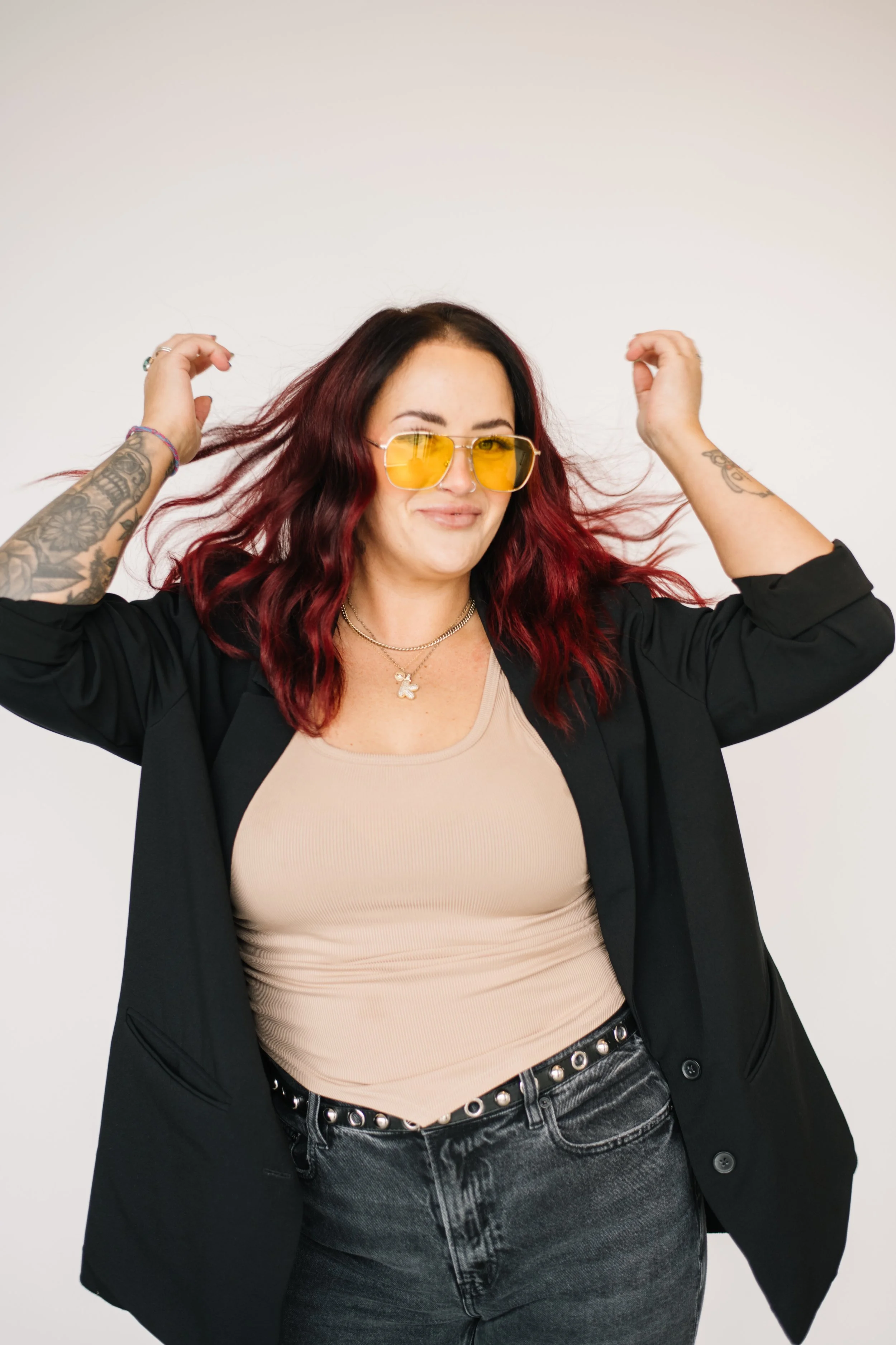 A woman with dark red hair wearing yellow sunglasses, a beige top, black jeans with silver studs, and a black blazer, striking a playful pose with her hands in her hair against a plain white background.