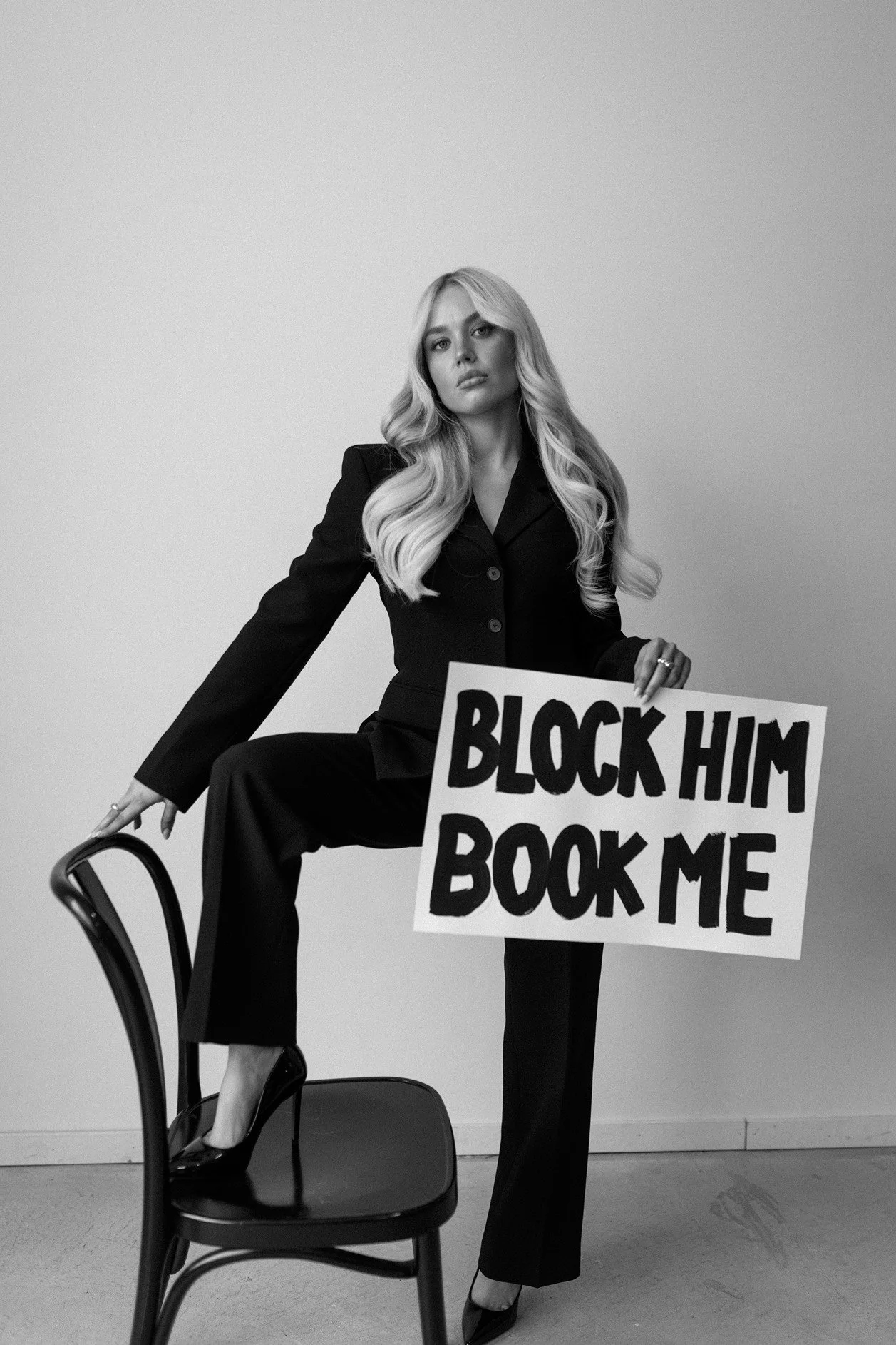 Black and white photograph of a woman in a suit sitting on a chair, holding a sign that says 'BLOCK HIM BOOK ME', with another chair in front of her and a plain wall background.