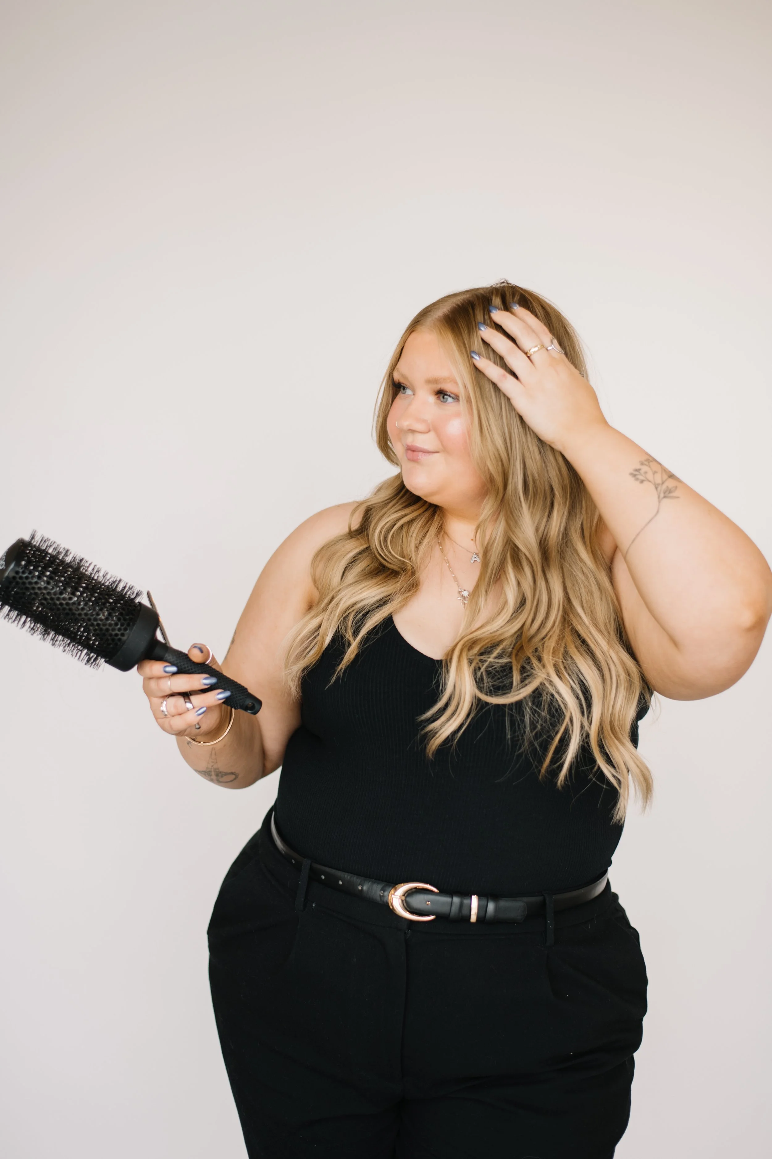 A woman with long blonde hair styled in waves, wearing a black sleeveless top, and holding a round hairbrush, is standing against a plain white background. She is touching her hair with her right hand, and has a tattoo on her right forearm, multiple rings on her fingers, and a necklace.