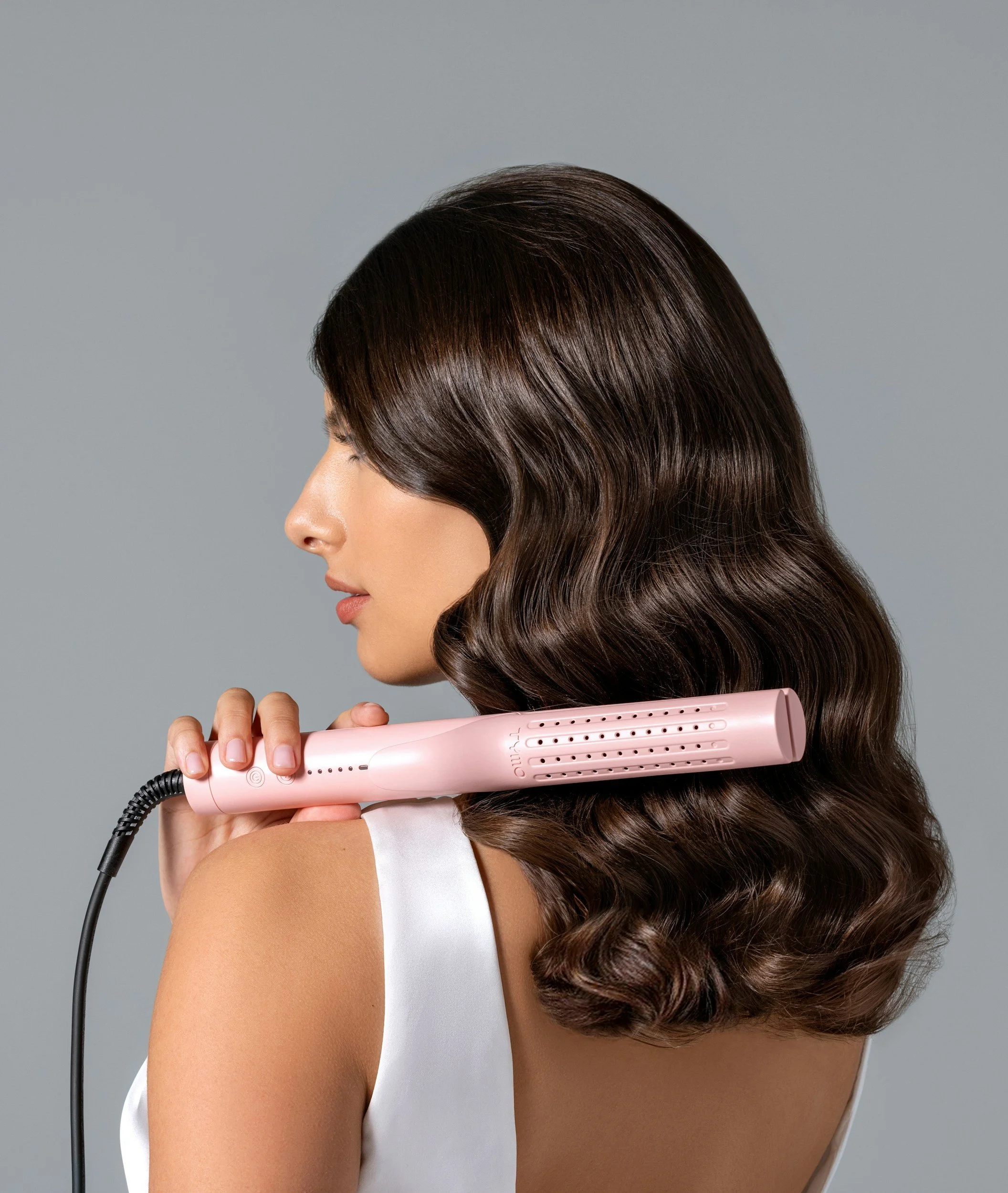 A woman with shiny brown hair holding a pink hair straightener to her shoulder.