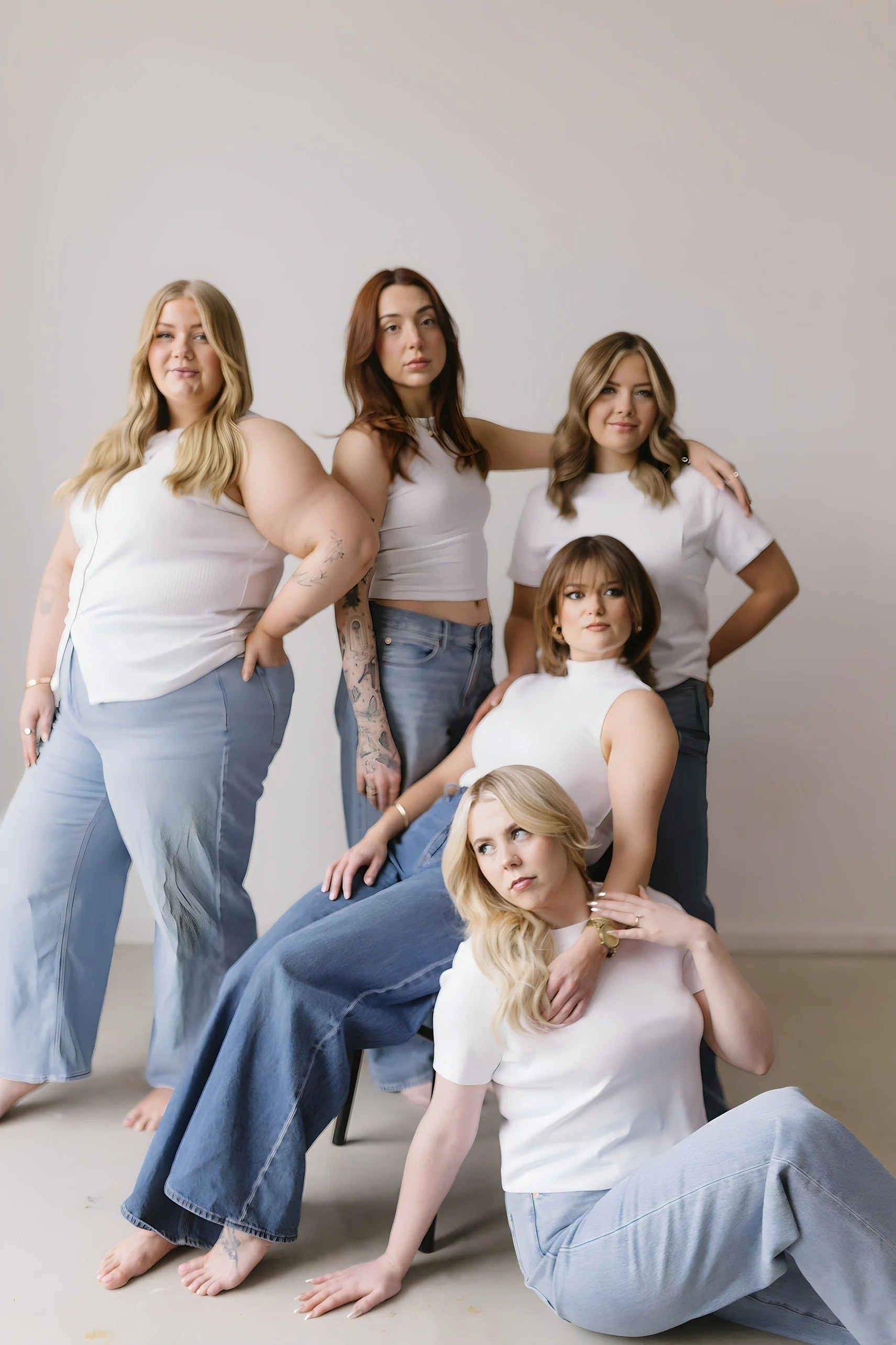 Six women with varying hair colors and styles posing together indoors against a plain background, dressed in casual white tops and blue jeans.