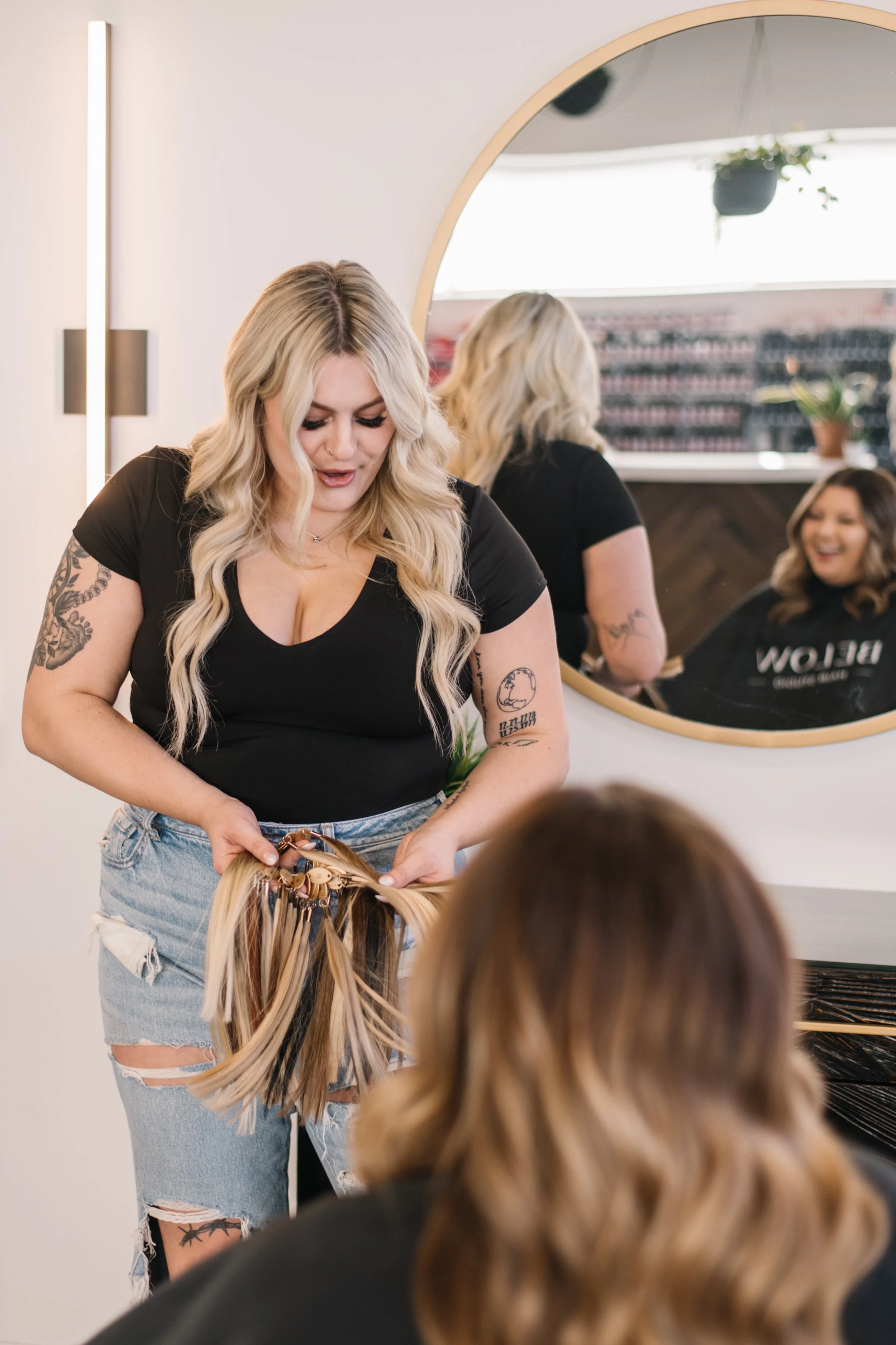 A woman with long blond hair and tattoos on her arms, wearing a black shirt and ripped jeans, is styling a client's hair with hair clips in a salon. There is a large round mirror behind her reflecting her and the salon interior, including a window with a hanging plant and shelves with products. Two other women with long hair are seated, one of whom is smiling.