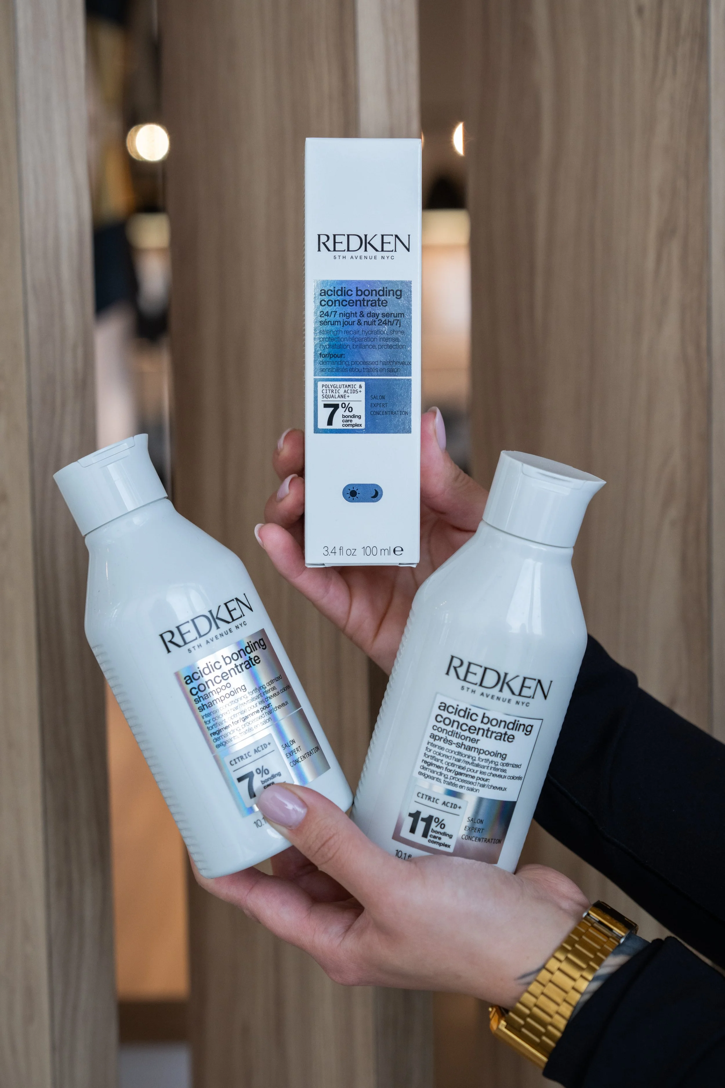 A person holding three bottles of Redken Acidic Bonding Concentrate hair care products, including shampoo and conditioner, against a wooden background.