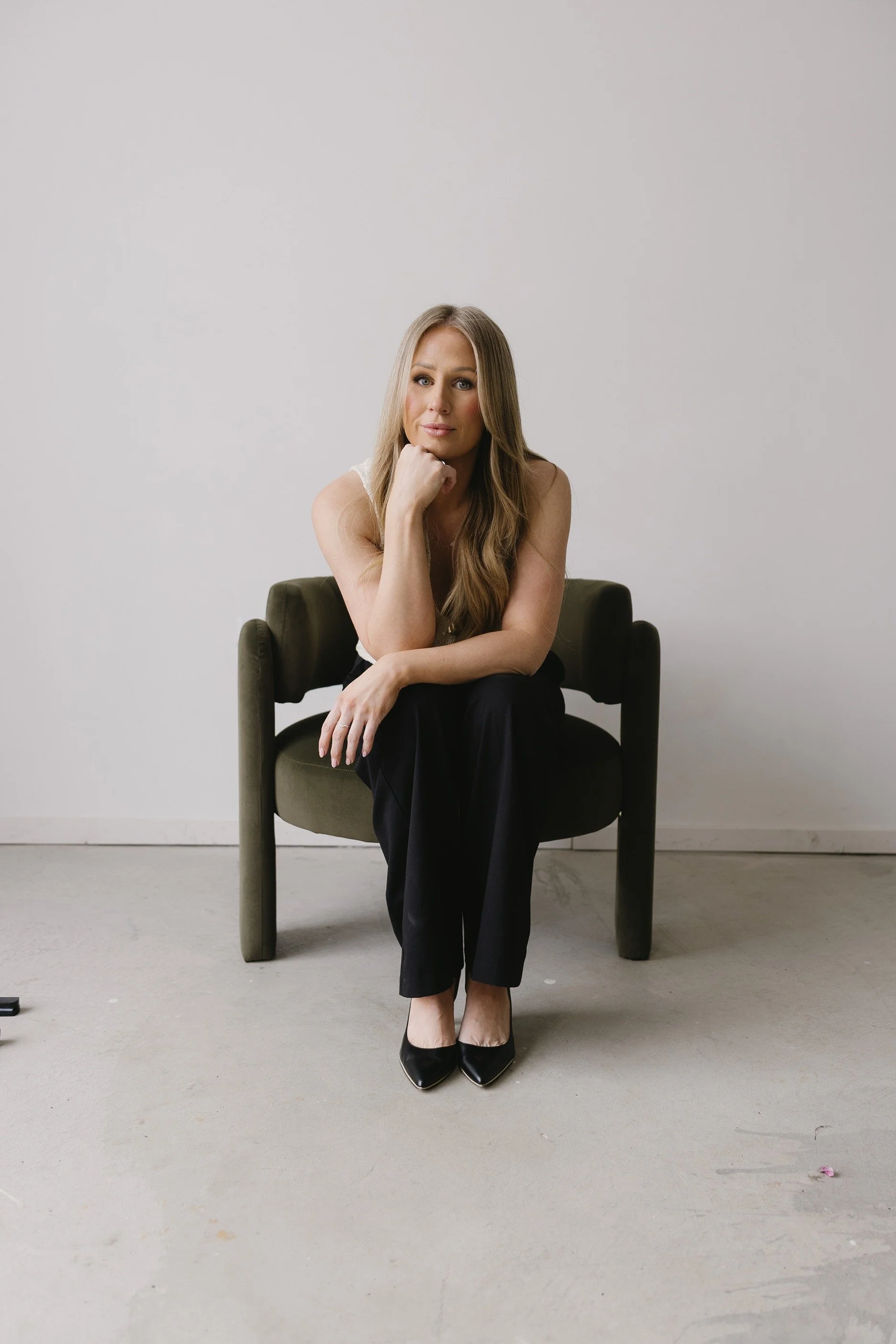 A woman with long, wavy blonde hair sitting on a dark green chair in front of a plain light gray wall, wearing a sleeveless white top, black pants, and black pointed shoes, with her chin resting on her left hand, looking directly at the camera.