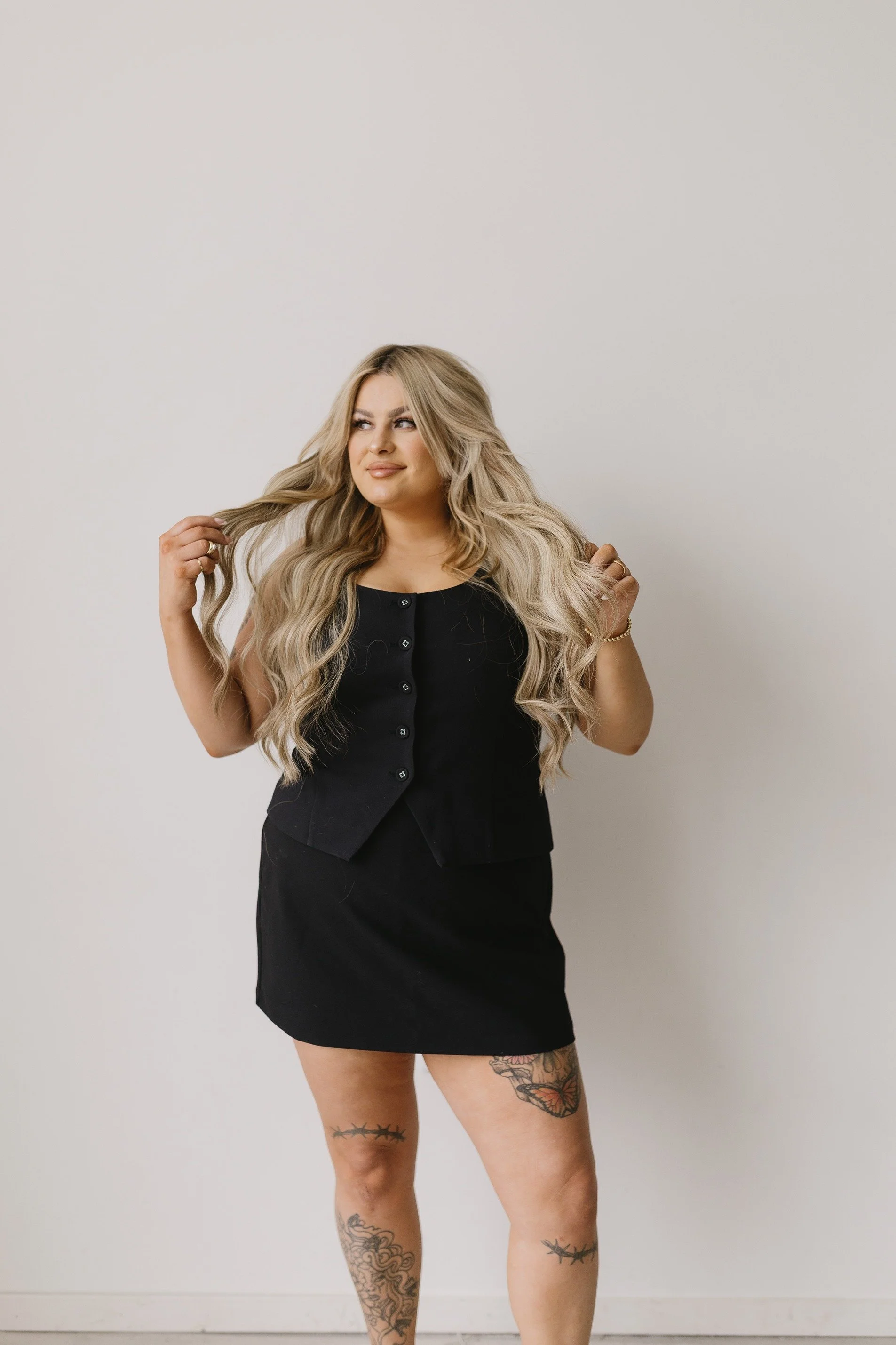 A woman with long, wavy blonde hair, wearing a black sleeveless button-up top and black skirt, stands against a plain white wall. She has tattoos on her thighs and is holding her hair with both hands.