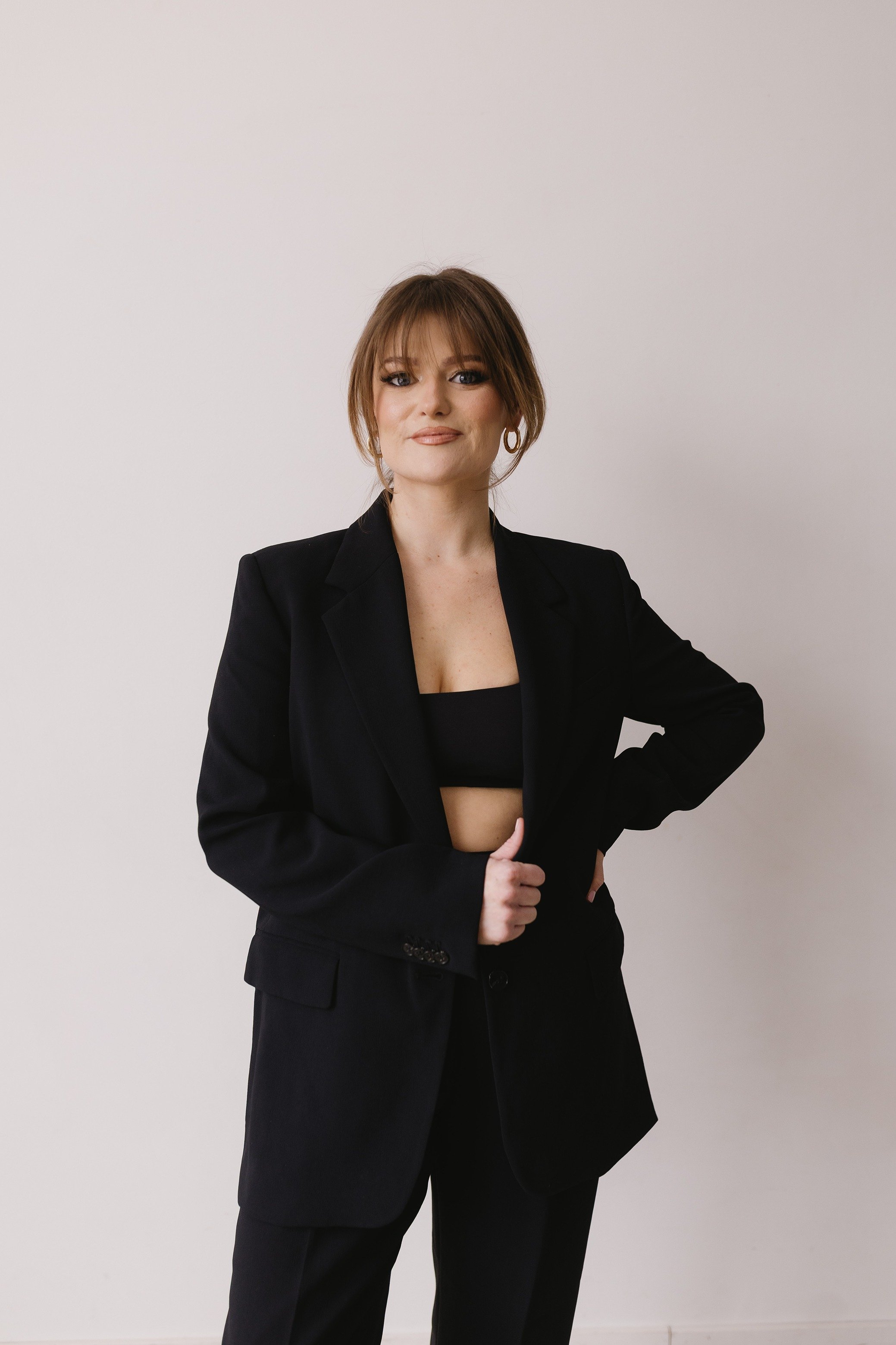 A woman with brown hair styled in a loose updo, wearing a black blazer and pants, standing against a plain white wall.