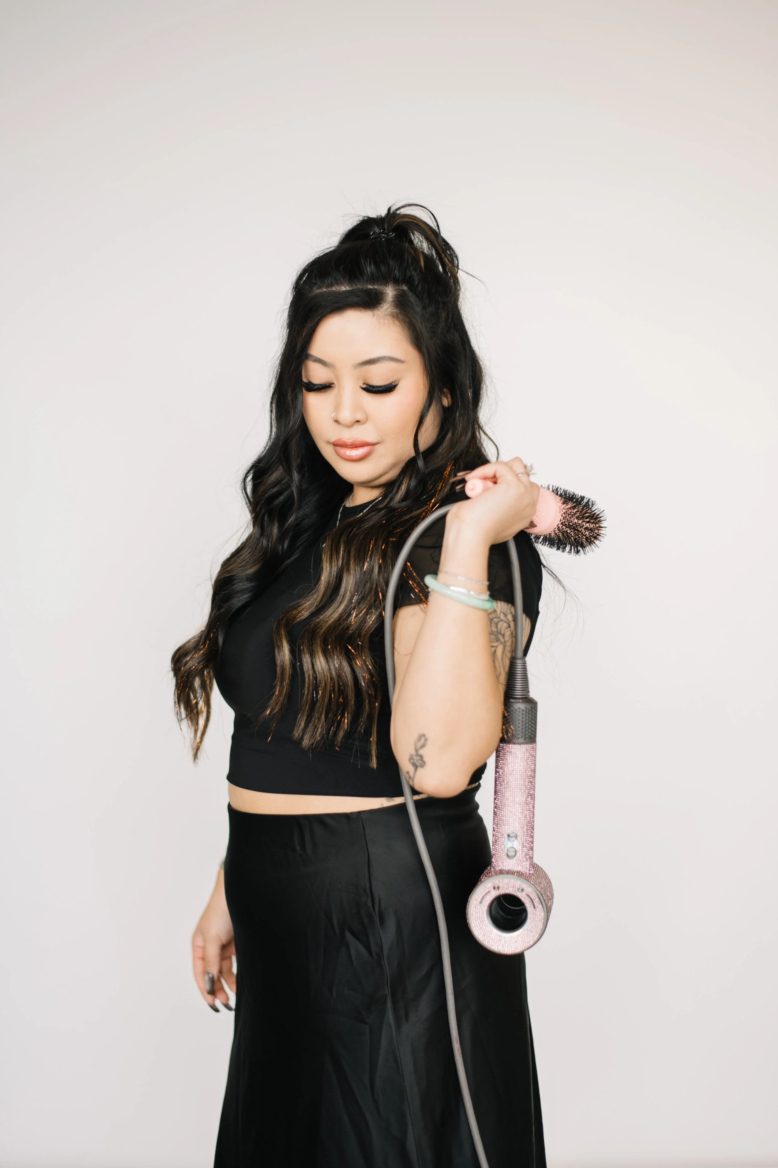 A woman with long dark hair styled in waves, wearing black makeup and a black crop top, holding a pink hairbrush connected to a pink hairdryer, standing against a plain white background.