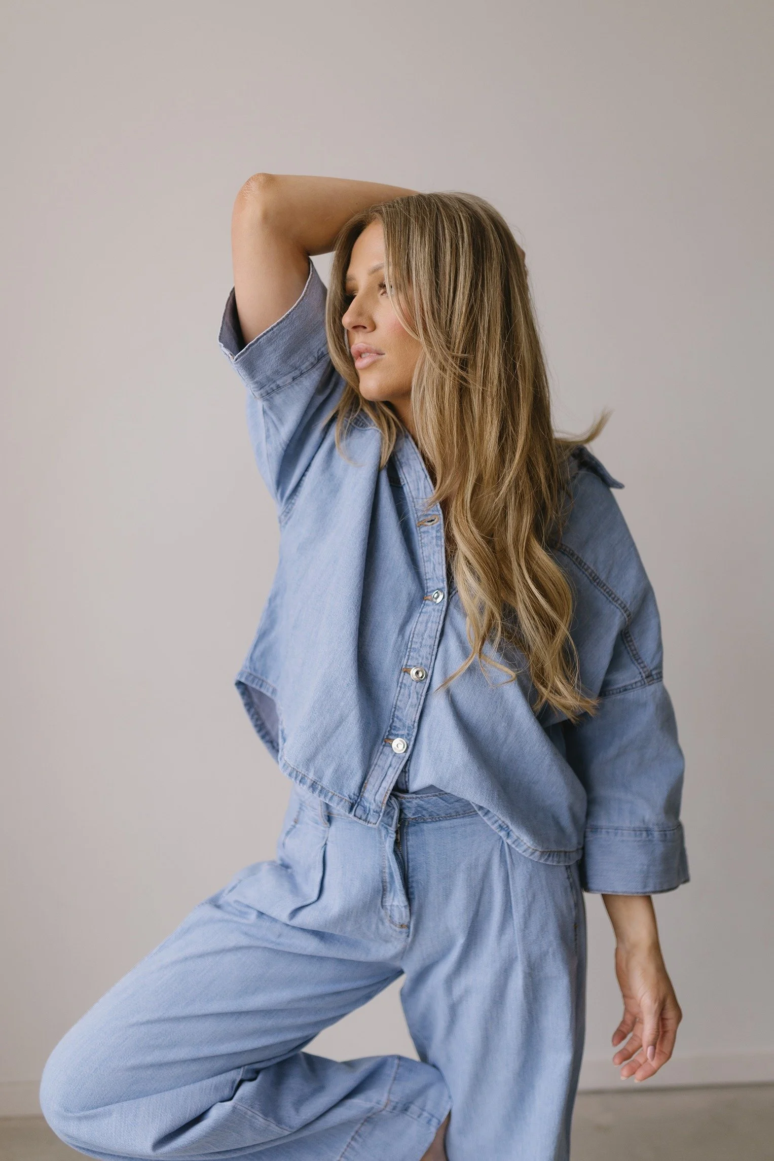 A woman with long, wavy hair wearing a light blue denim jacket and matching pants, sitting on the floor with one knee up and her hand resting on her knee, against a plain light background.