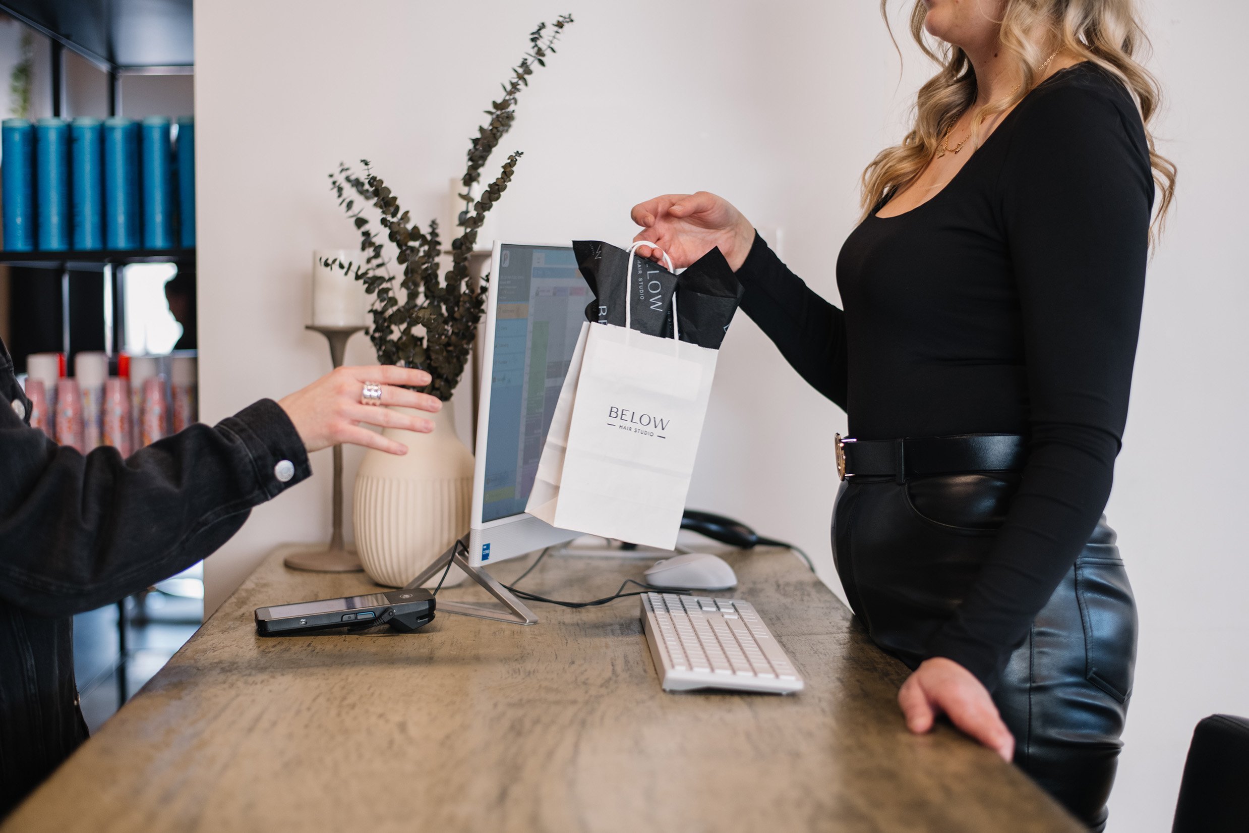 A woman with long blonde hair wearing a black top and leather skirt is handing a paper shopping bag to another person at a reception desk. The person receiving the bag is wearing a black leather jacket. The reception desk has a computer, keyboard, mouse, and a smartphone. In the background, there is a large beige vase with decorative branch, books, and shelves.