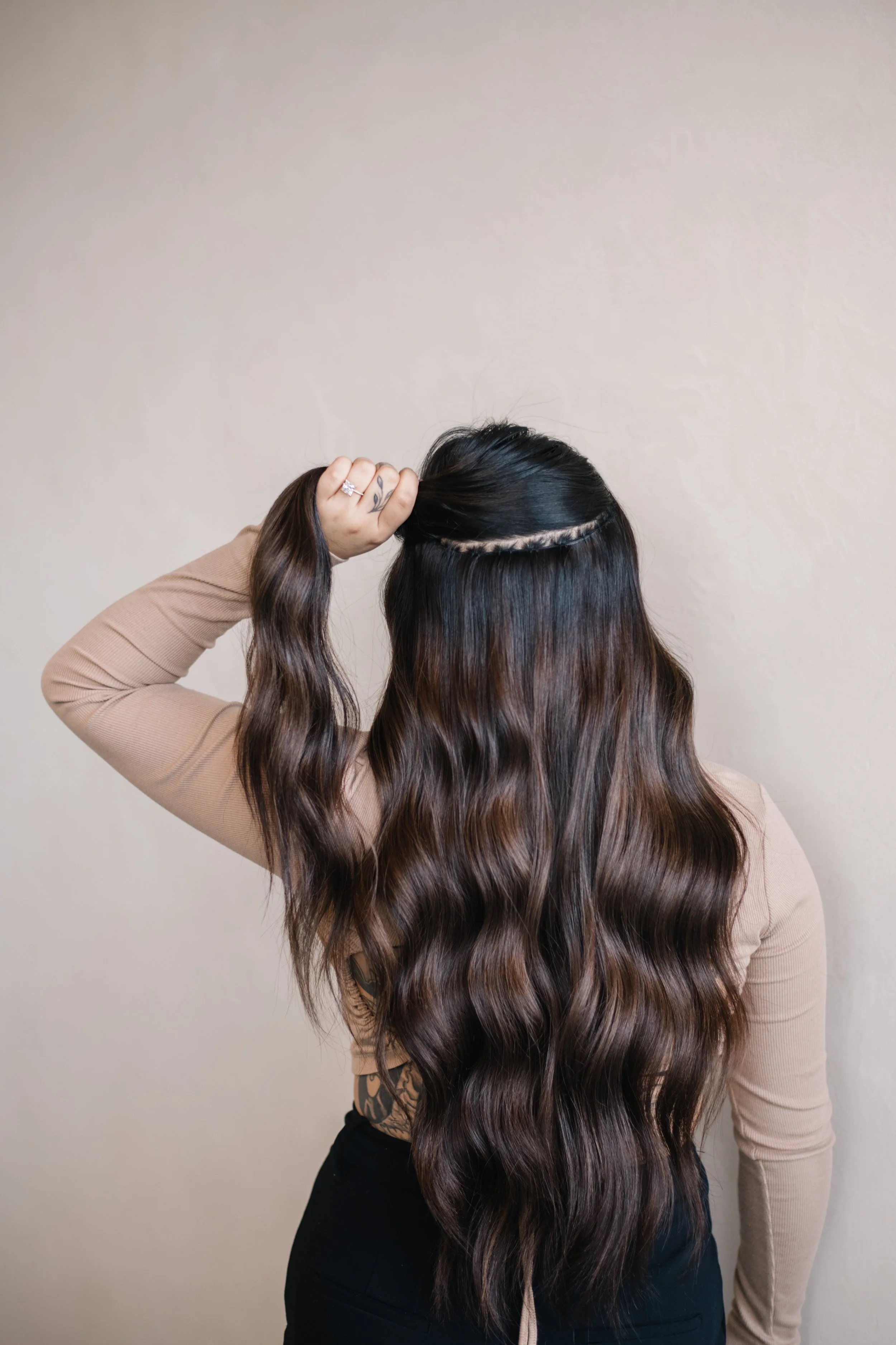 A woman with long wavy brown hair, wearing a tan long-sleeve shirt, is seen from behind, against a beige wall. She is holding her hair up with her left hand, showing her patterned tattooed arm, which has a diamond ring on her finger.