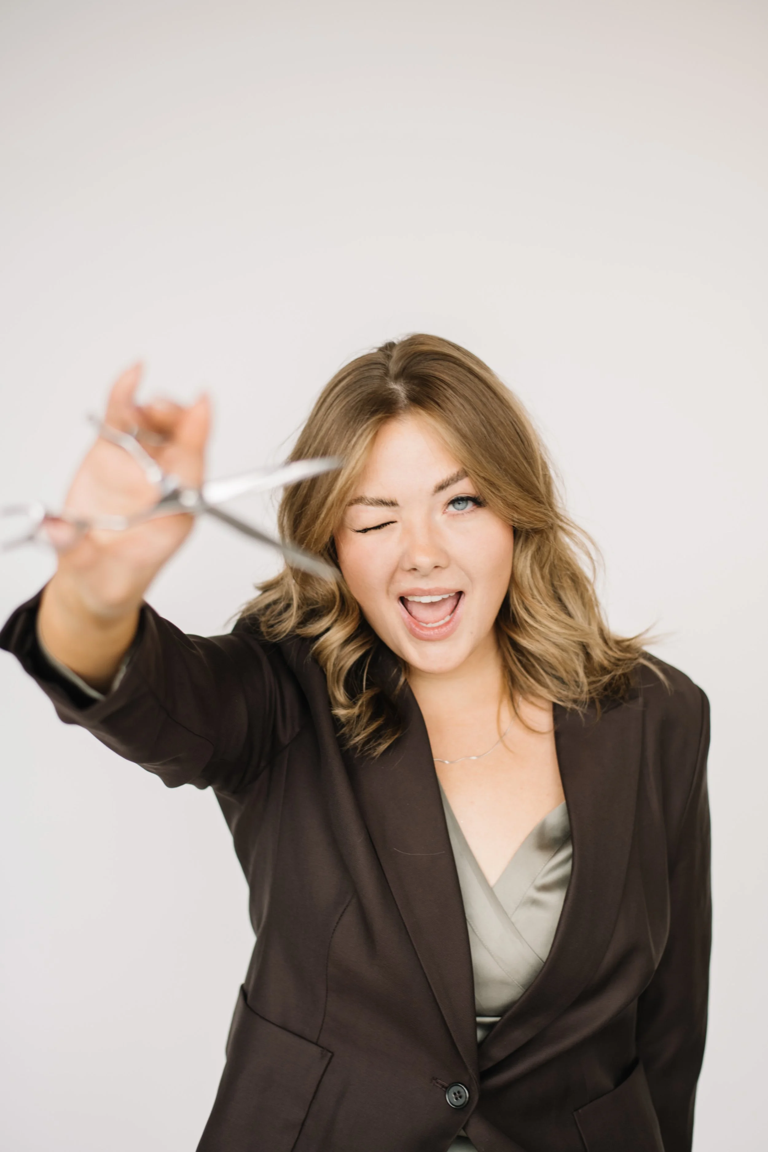 Woman with wavy blonde hair and blue eyes, holding a pair of scissors in front, wearing a black blazer over a satin top, smiling and winking against a plain white background.