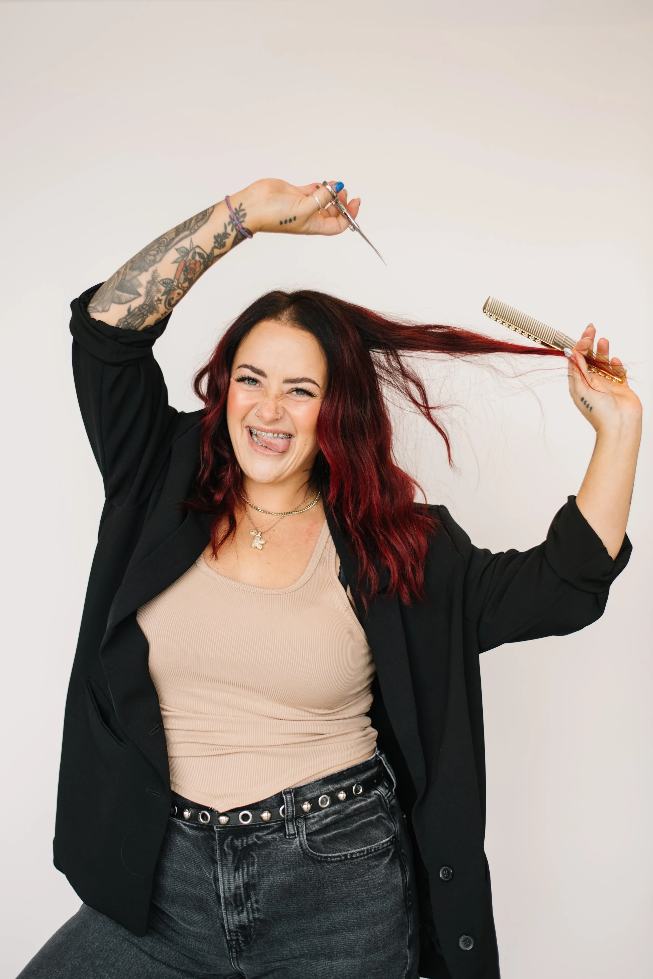 A woman with red hair and tattoos on her arm, wearing a black blazer and beige top, holding a comb in one hand and scissors in the other, making a playful face with her tongue out.
