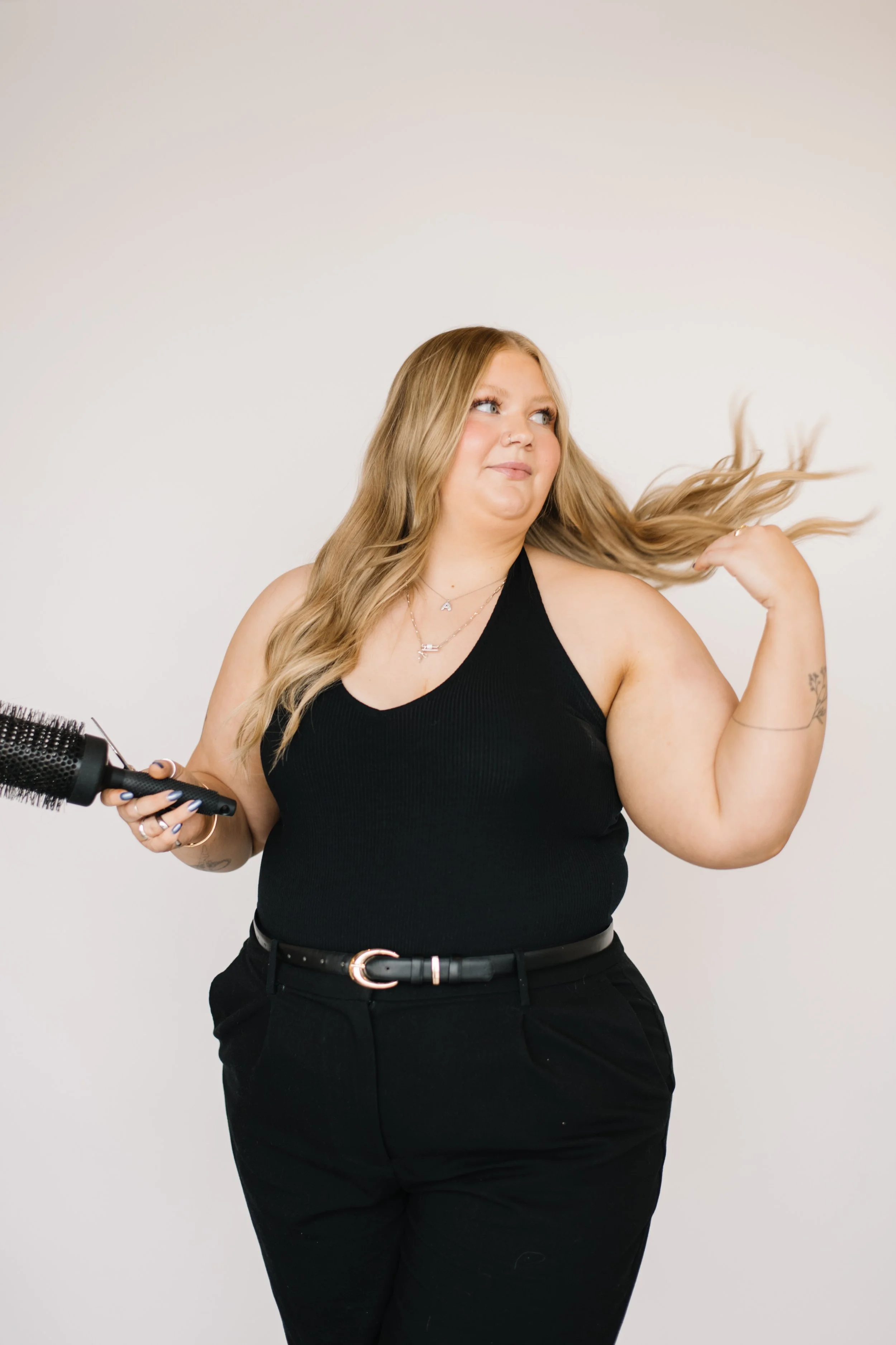 A woman with long blonde hair, wearing a black tank top and black pants, holding a round hairbrush in her right hand, adjusting her hair with her left hand, standing against a plain light gray background.