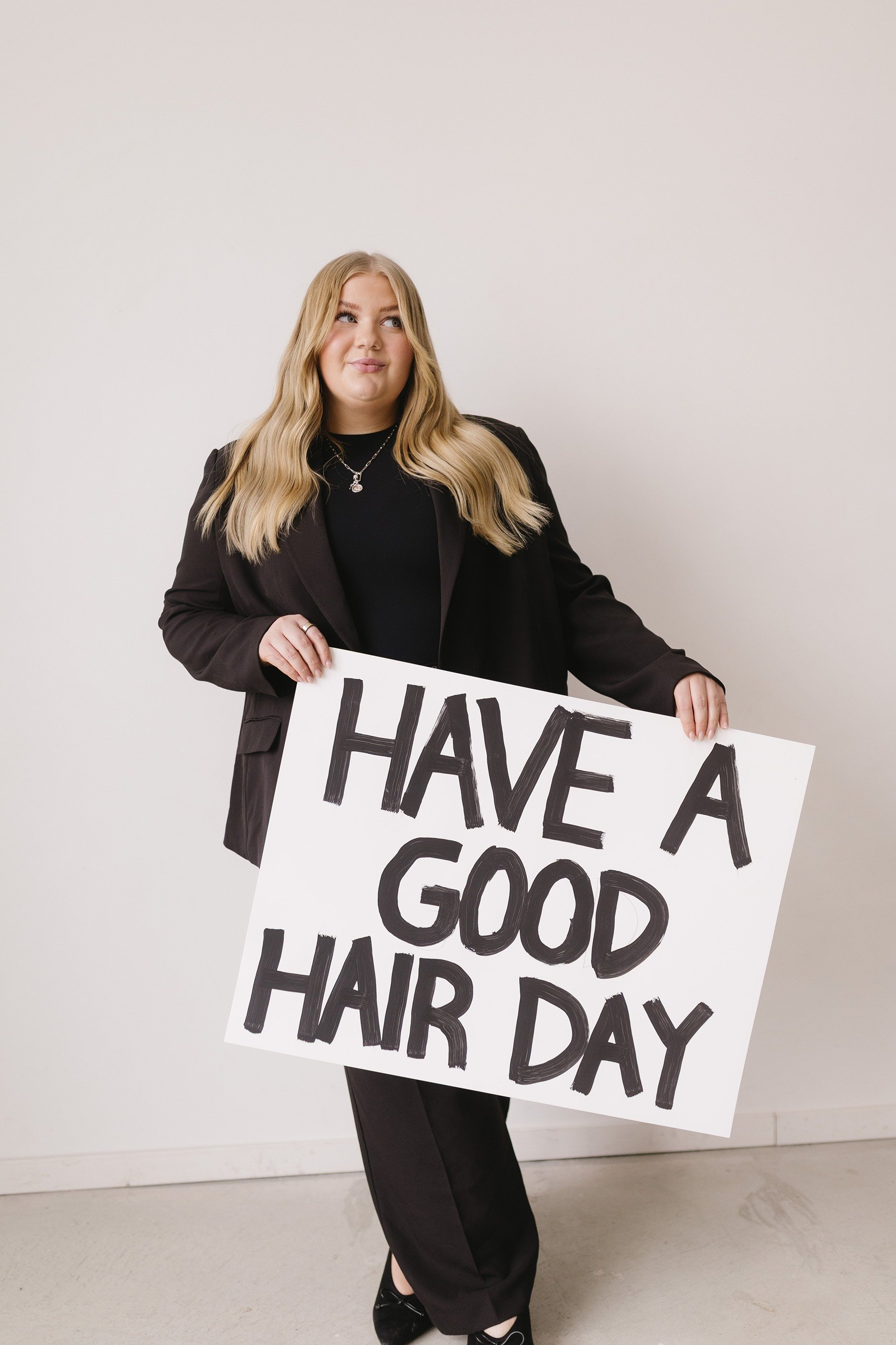 A woman with long blonde hair wearing a black blazer and dress holding a large sign that reads "HAVE A GOOD HAIR DAY." She is standing against a plain white background.