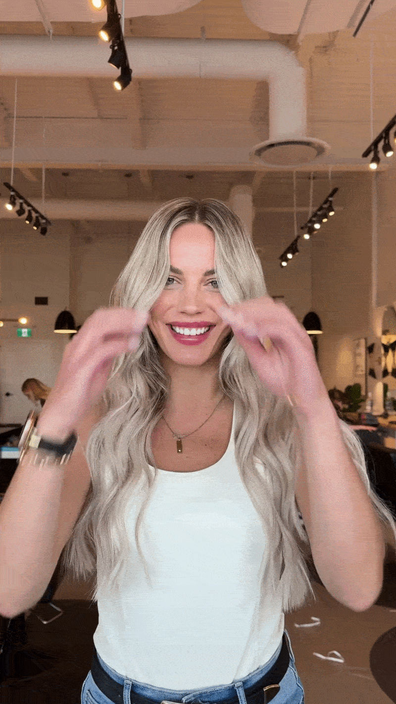 A smiling woman with long blonde hair wearing a white tank top and blue jeans, standing in a modern indoor space with wood ceilings, black track lighting, and other people in the background.