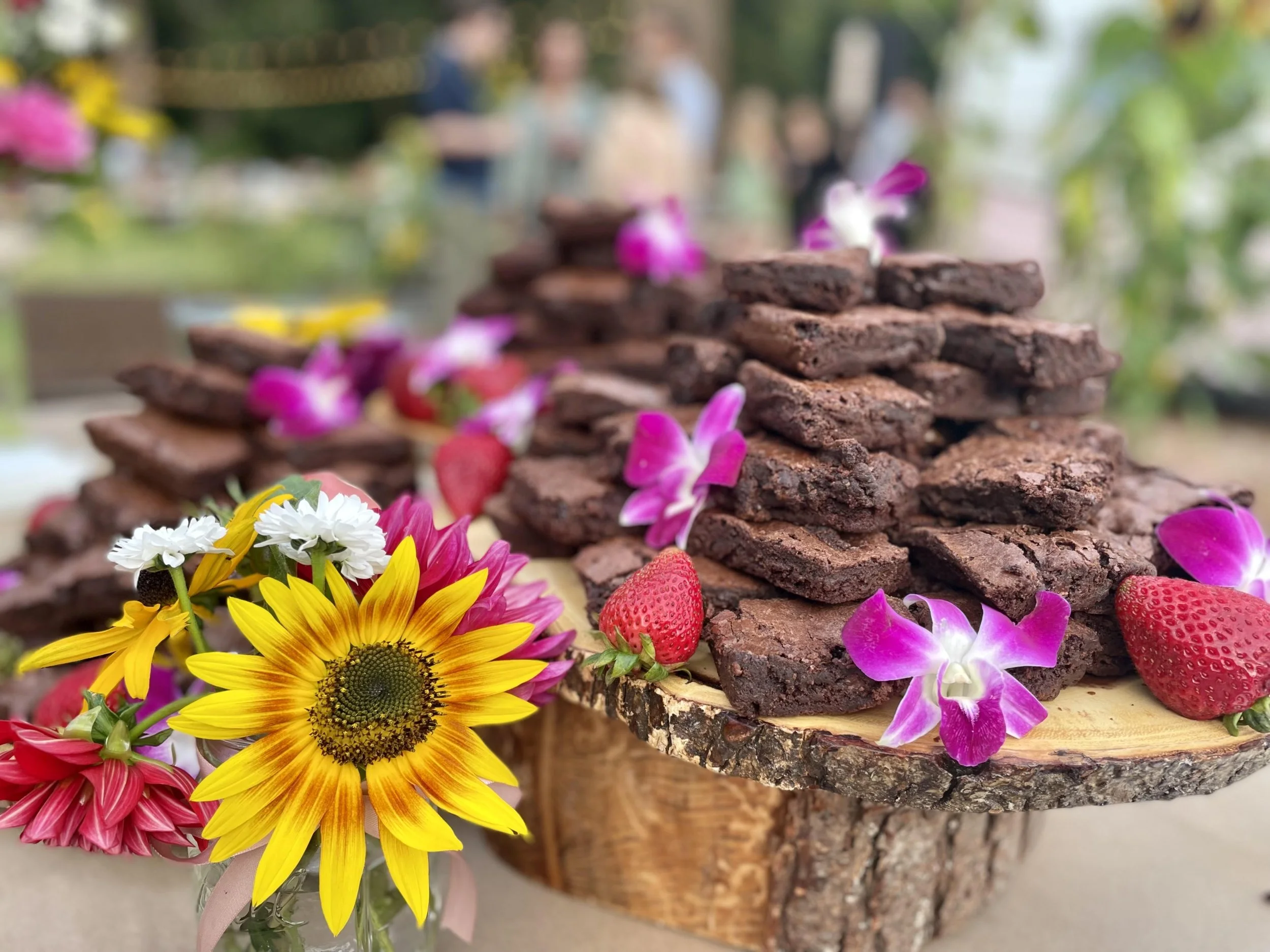 A wooden table displays a cake stand made from a tree stump, with stacked brownies decorated with pink and purple orchids and strawberries. A bouquet of colorful flowers, including sunflowers, daisies, and pink and red blooms, is nearby. In the blurr