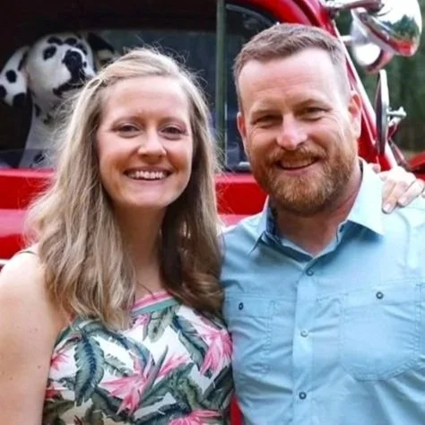 A woman and a man smiling in front of a red fire truck with a panda mascot in the background.