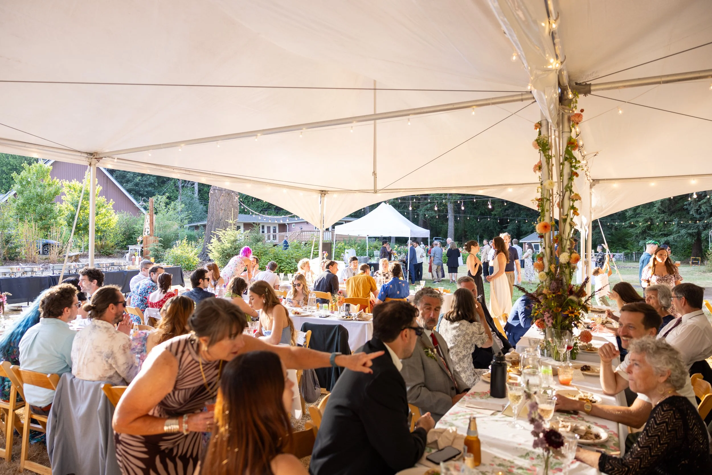 A large outdoor gathering under a white tent at dusk, with many people seated at long tables decorated with flowers, dining and socializing.