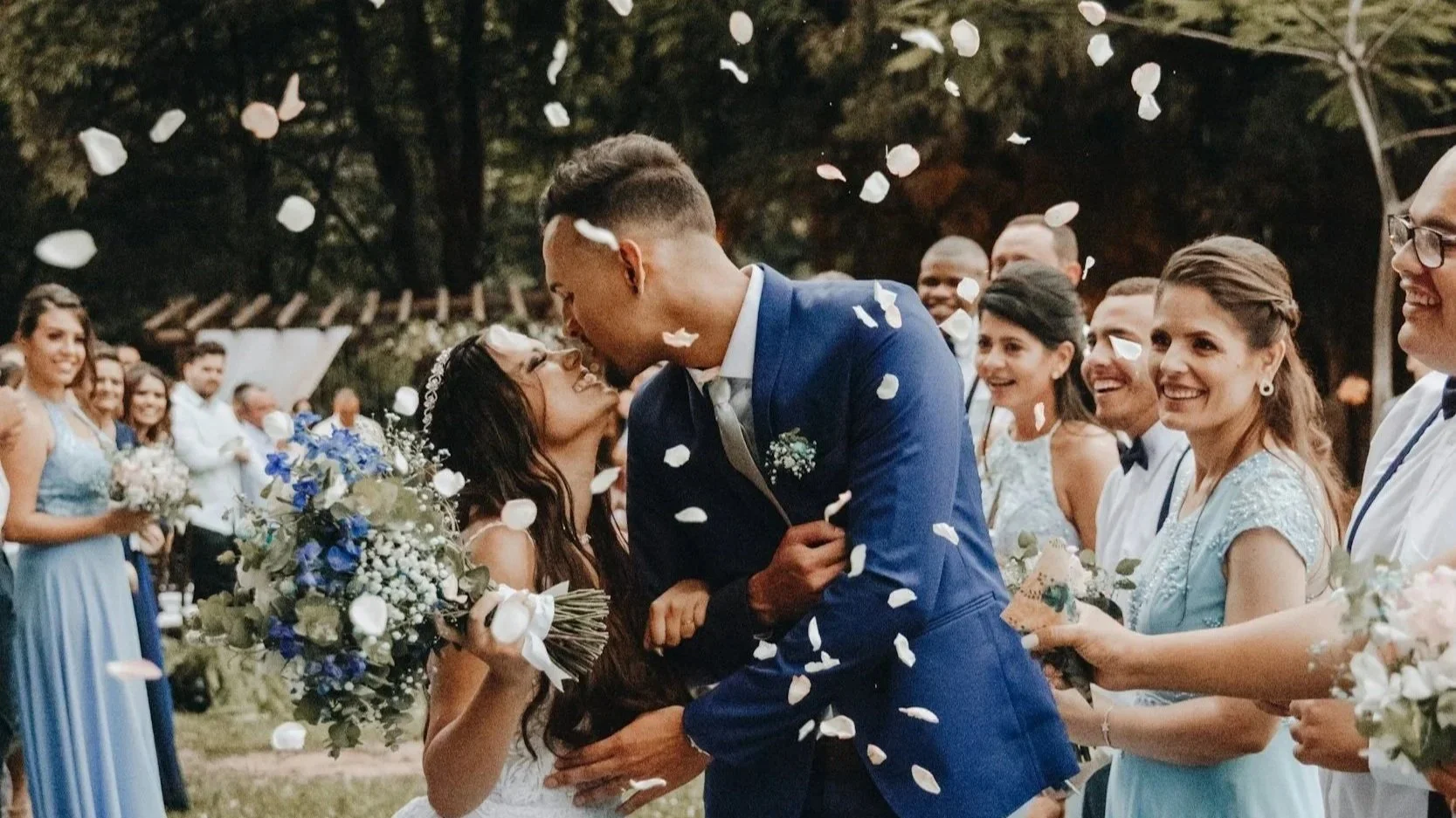 A couple getting married stand under a wooden floral arch in an outdoor setting, with an officiant holding a microphone and reading from a paper, while guests watch, surrounded by lush greenery.