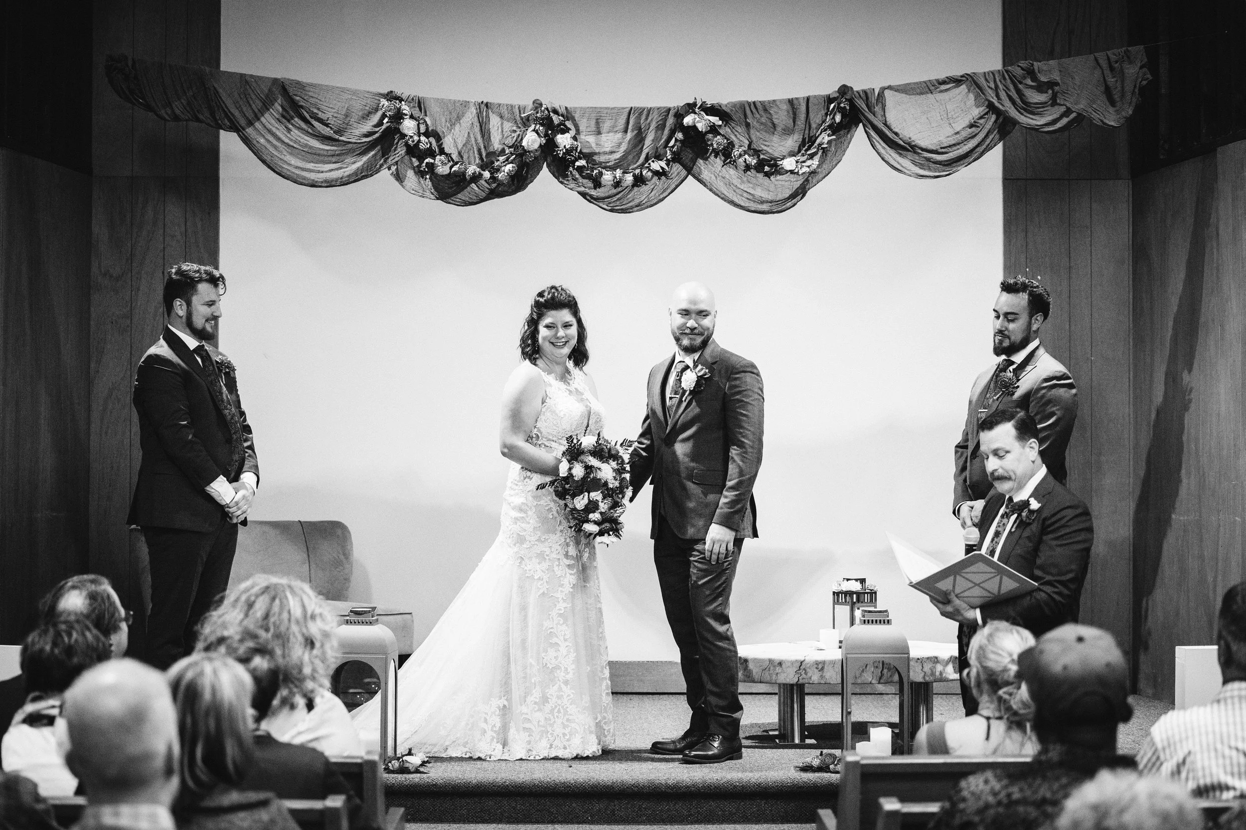 A smiling man and woman stand in front of a church with a modern A-frame design surrounded by greenery and trees.