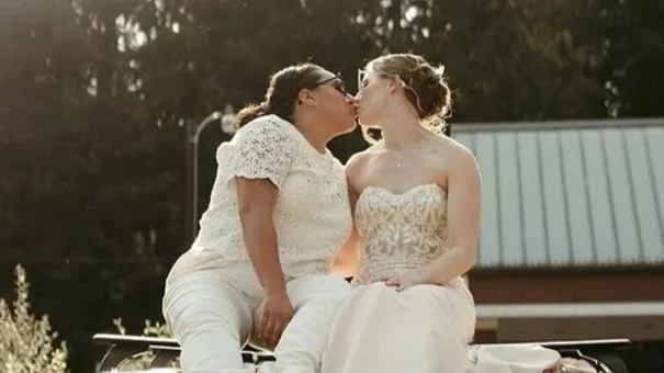 A couple getting married stand under a wooden floral arch in an outdoor setting, with an officiant holding a microphone and reading from a paper, while guests watch, surrounded by lush greenery.