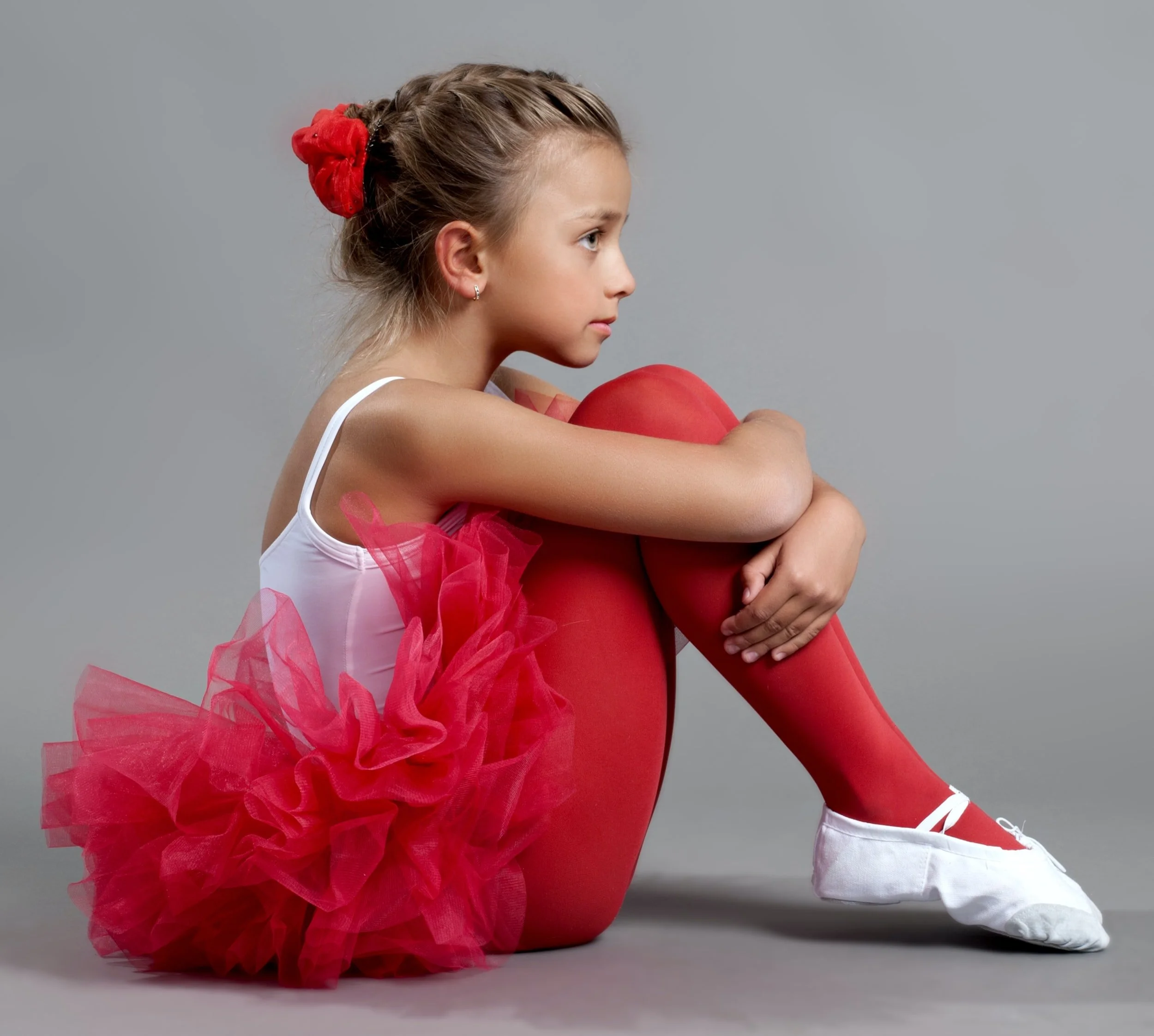 A young girl dressed as a ballerina in a pink tutu, pink tights, white ballet shoes, and white leotard, sitting with her arms wrapped around her knees, against a gray background.