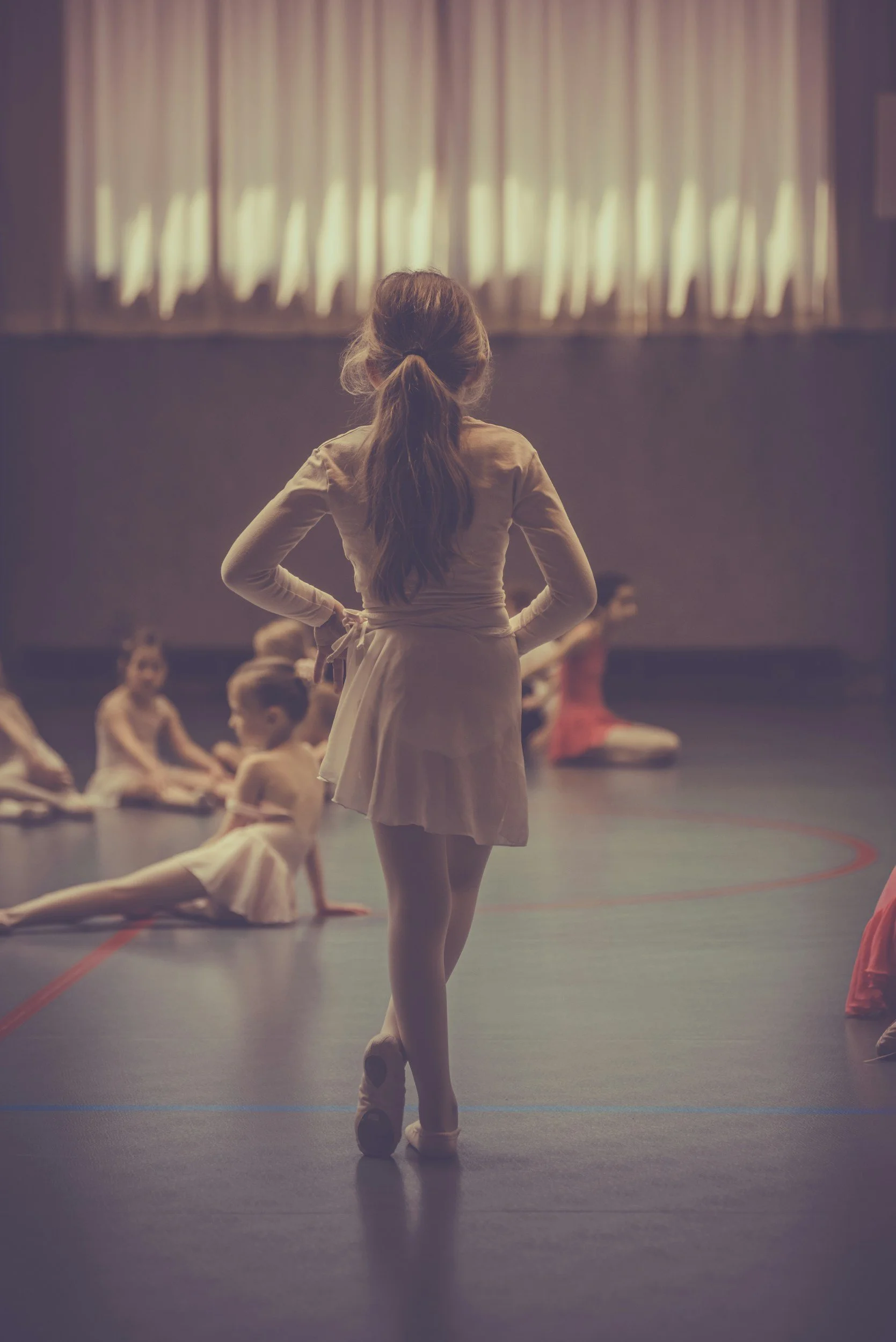 A girl standing on stage in a ballet costume, facing away, with other children in ballet costumes seated and lying on the stage floor in the background, and a curtain with stage lights above.