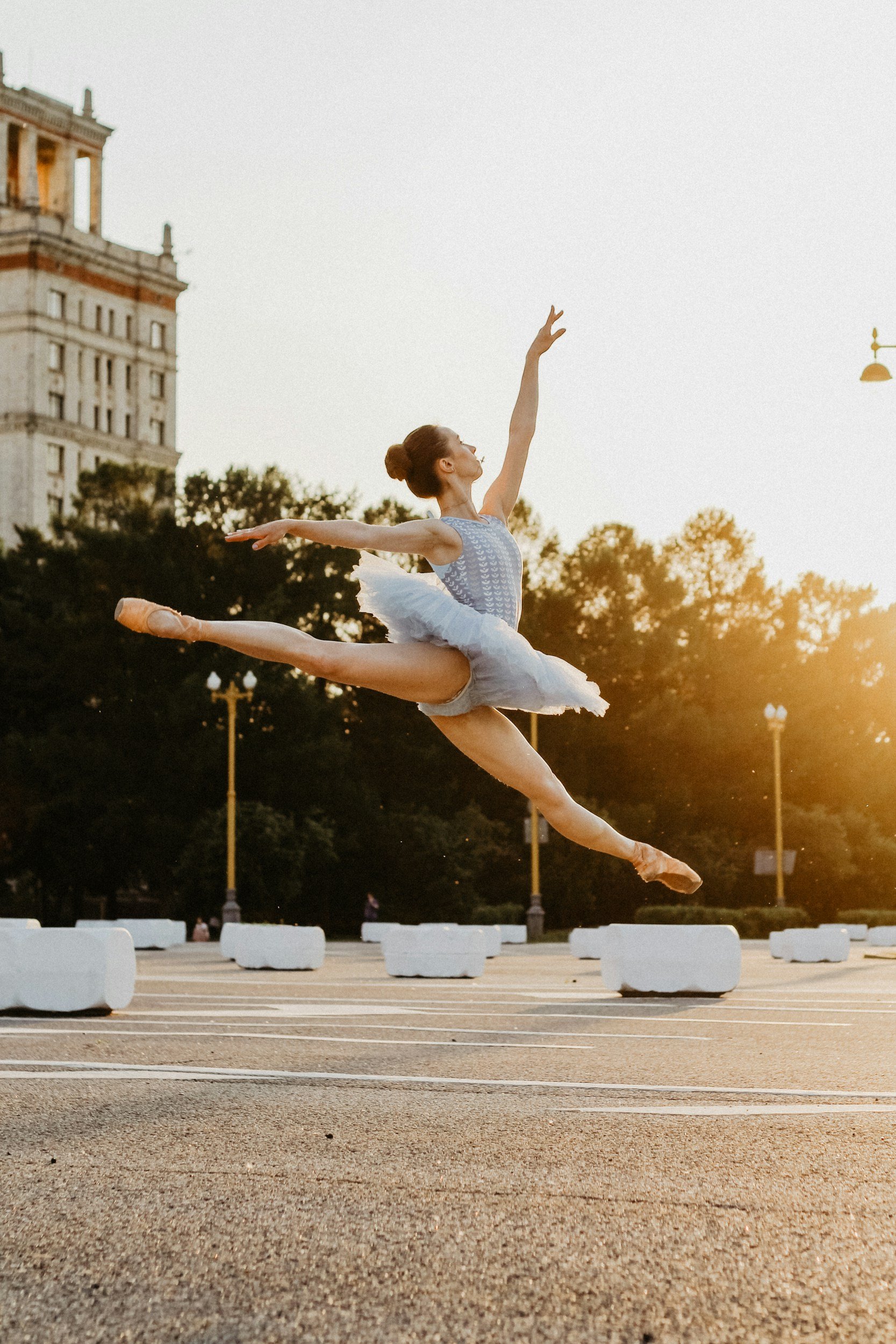 A ballet dancer performing a leap during sunset in an open urban space with white barriers and trees in the background.