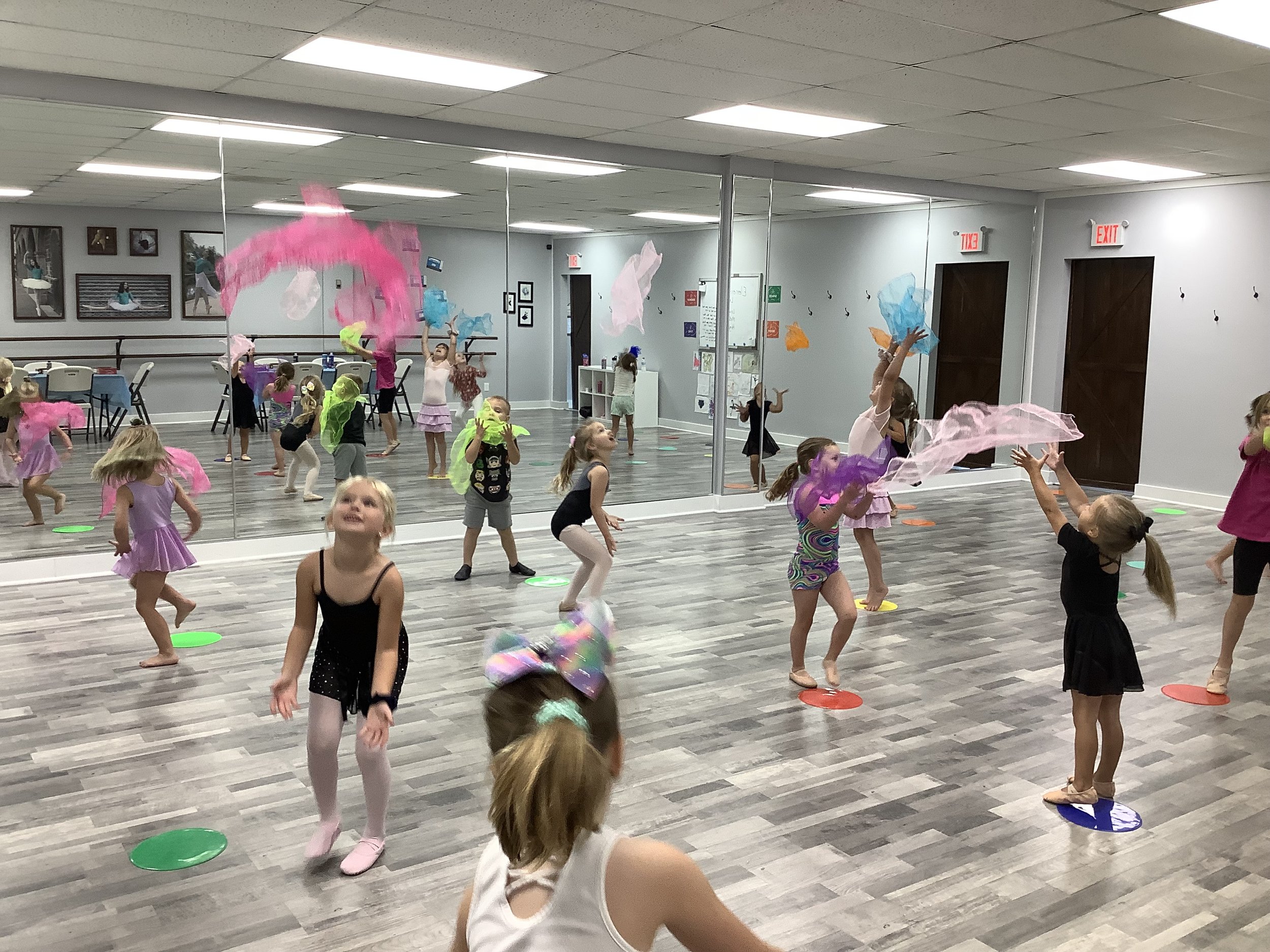 Young children participating in a dance or movement class in a spacious room with a mirrored wall, colorful scarves, and hoops on the floor.