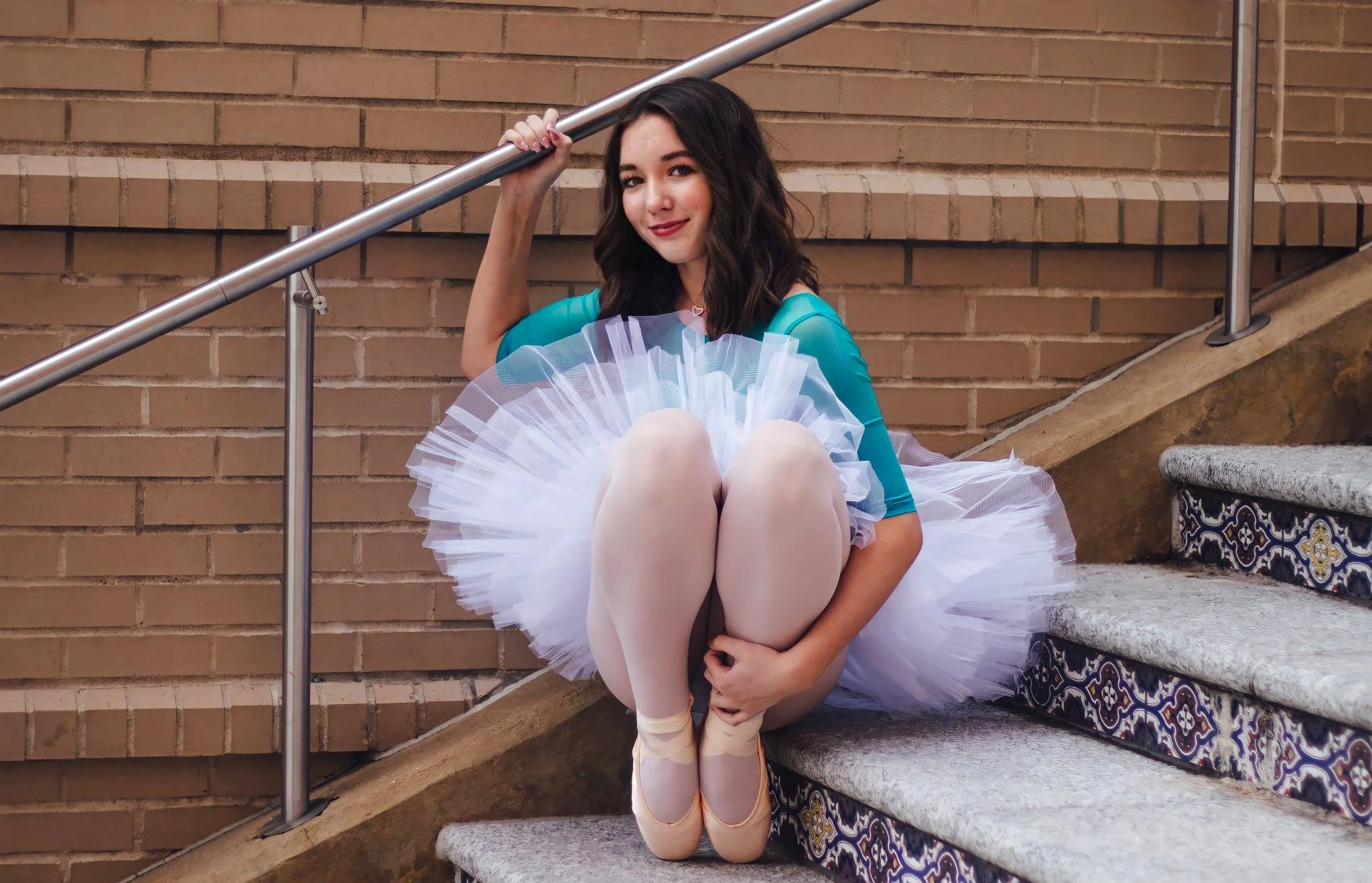 A young woman dressed as a ballerina sitting on steps against a brick wall, holding a tutu in her lap, with her legs crossed and a gentle smile.