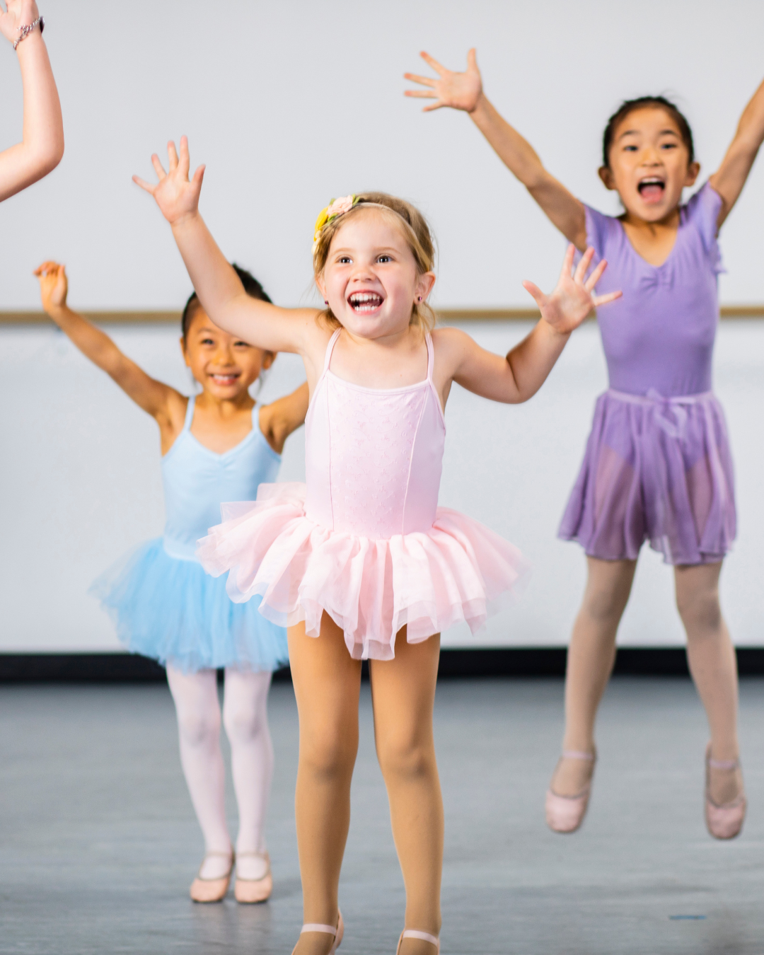 Three young girls in colorful ballet tutus and tights performing ballet moves in a dance studio.