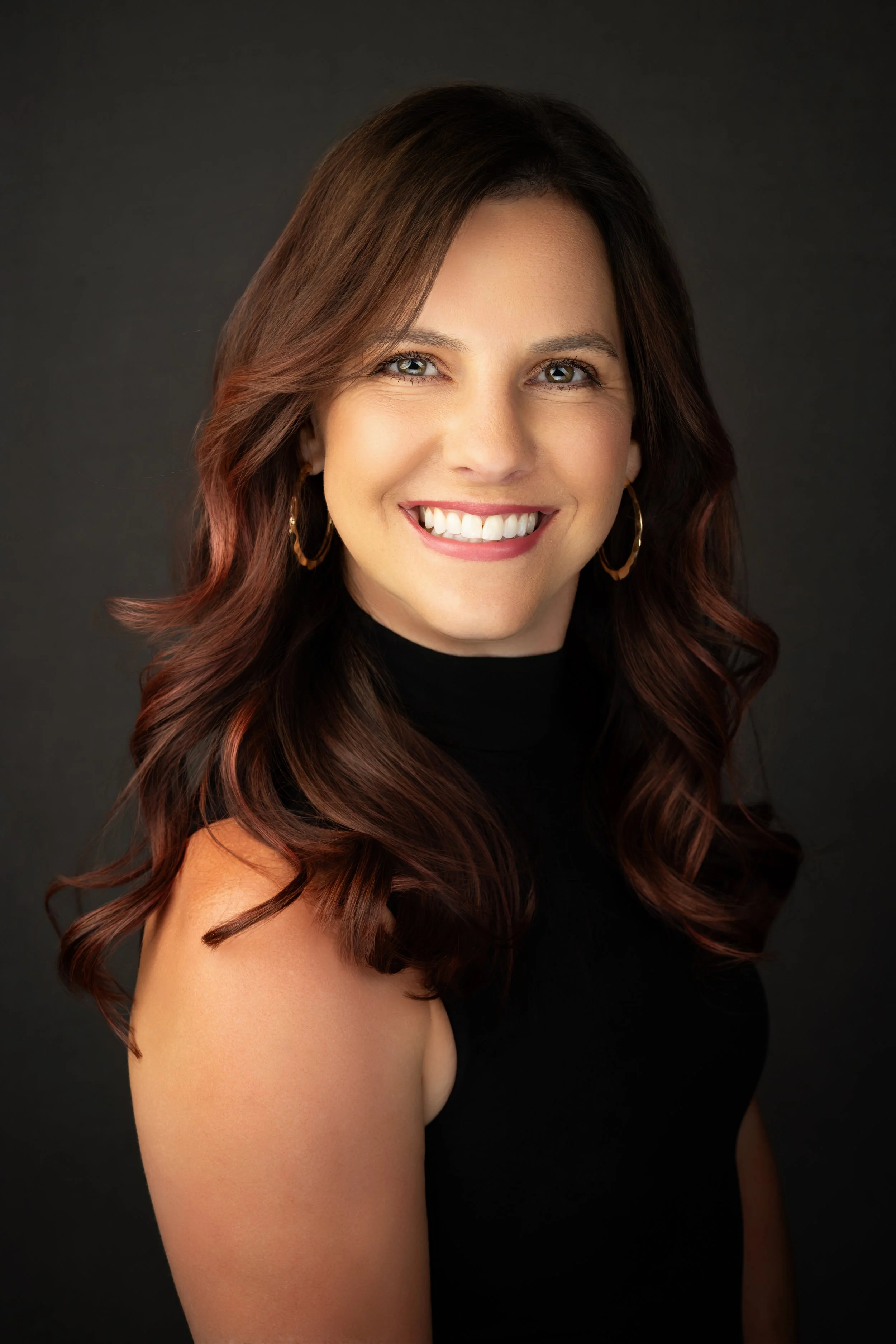 Headshot of a smiling woman with long wavy brown hair, wearing earrings and a black top, against a dark background.