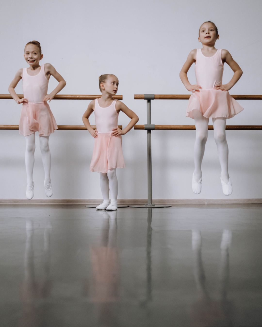 Three young girls in ballet costumes practicing at a dance studio, with two of them mid-air and one standing on the floor.